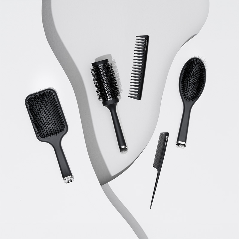 En samling svarta GHD-hårborstar och kammar är stilfullt arrangerade på en vit bakgrund med en böljande form. Designen är elegant och modern, med fokus på detaljer och funktionalitet.