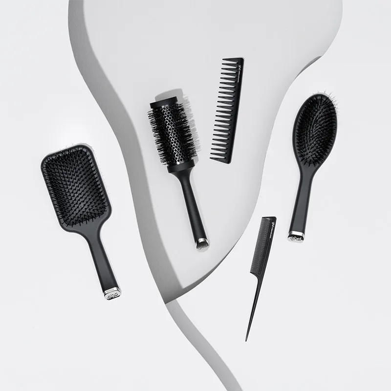 En samling svarta GHD-hårborstar och kammar är stilfullt arrangerade på en vit bakgrund med en böljande form. Designen är elegant och modern, med fokus på detaljer och funktionalitet.