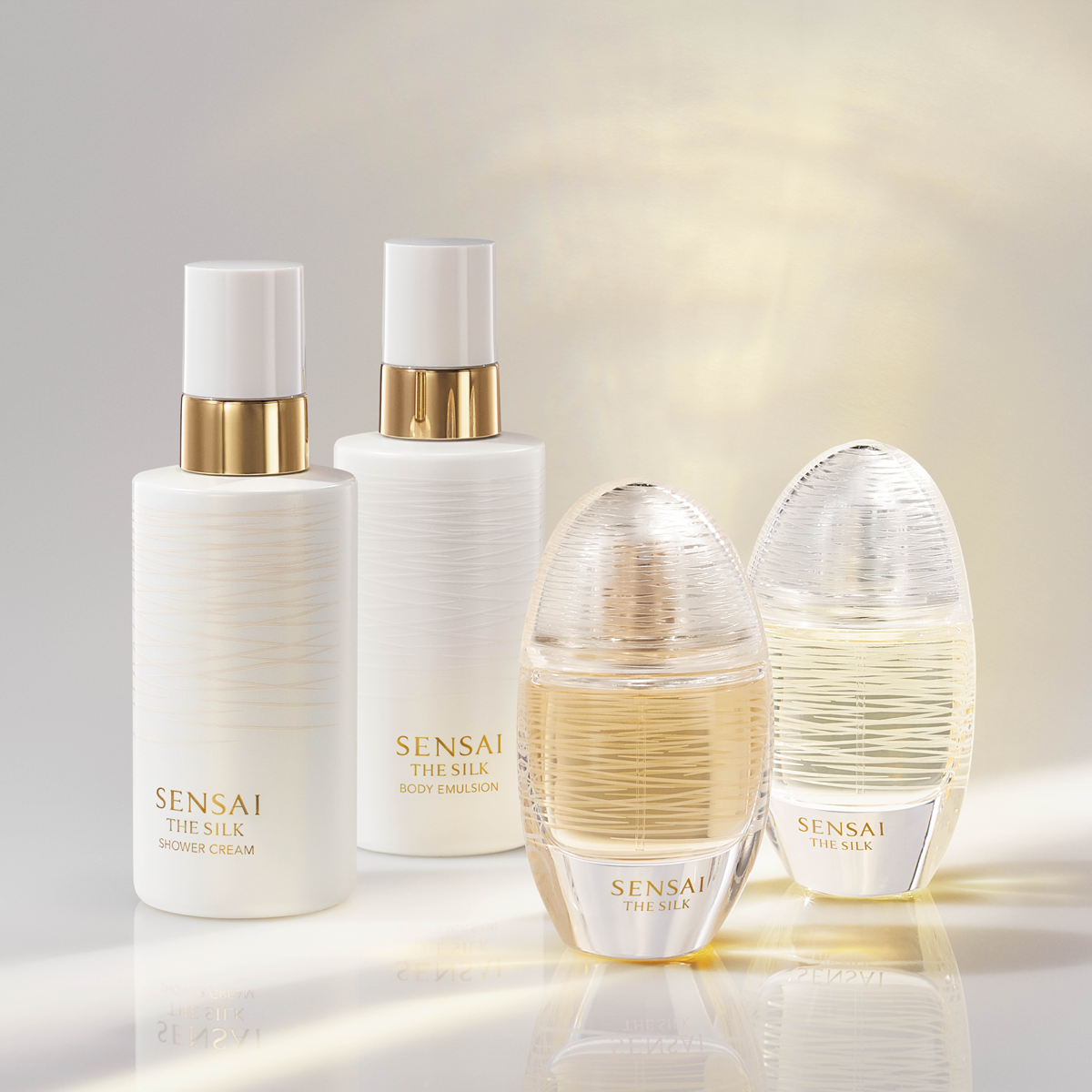 Neljä SENSAI The Silk -tuotetta esillä vaaleaa taustaa vasten: Shower Cream, Body Emulsion ja kaksi The Silk -sarjan hajuvesipulloa. Tuotteissa on valkoinen ja läpinäkyvä design kullanvärisillä yksityiskohdilla, jotka luovat ylellisen vaikutelman parfyymeista, body lotion -tuotteista ja vartalonhoidosta.