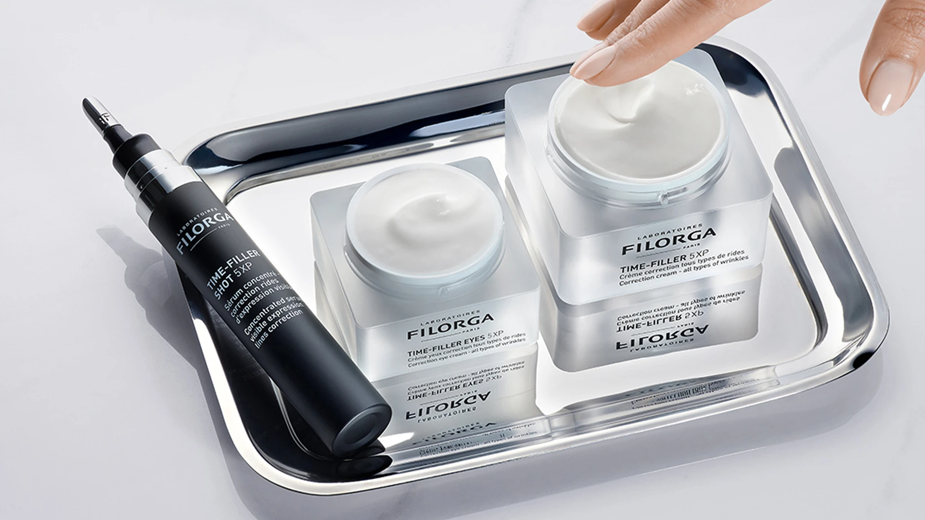 En hånd berører en krukke med Filorga "TIME-FILLER 5XP" krem, som står på et sølvbrett sammen med en mindre krukke og en tube med "TIME-FILLER EYES 5XP".