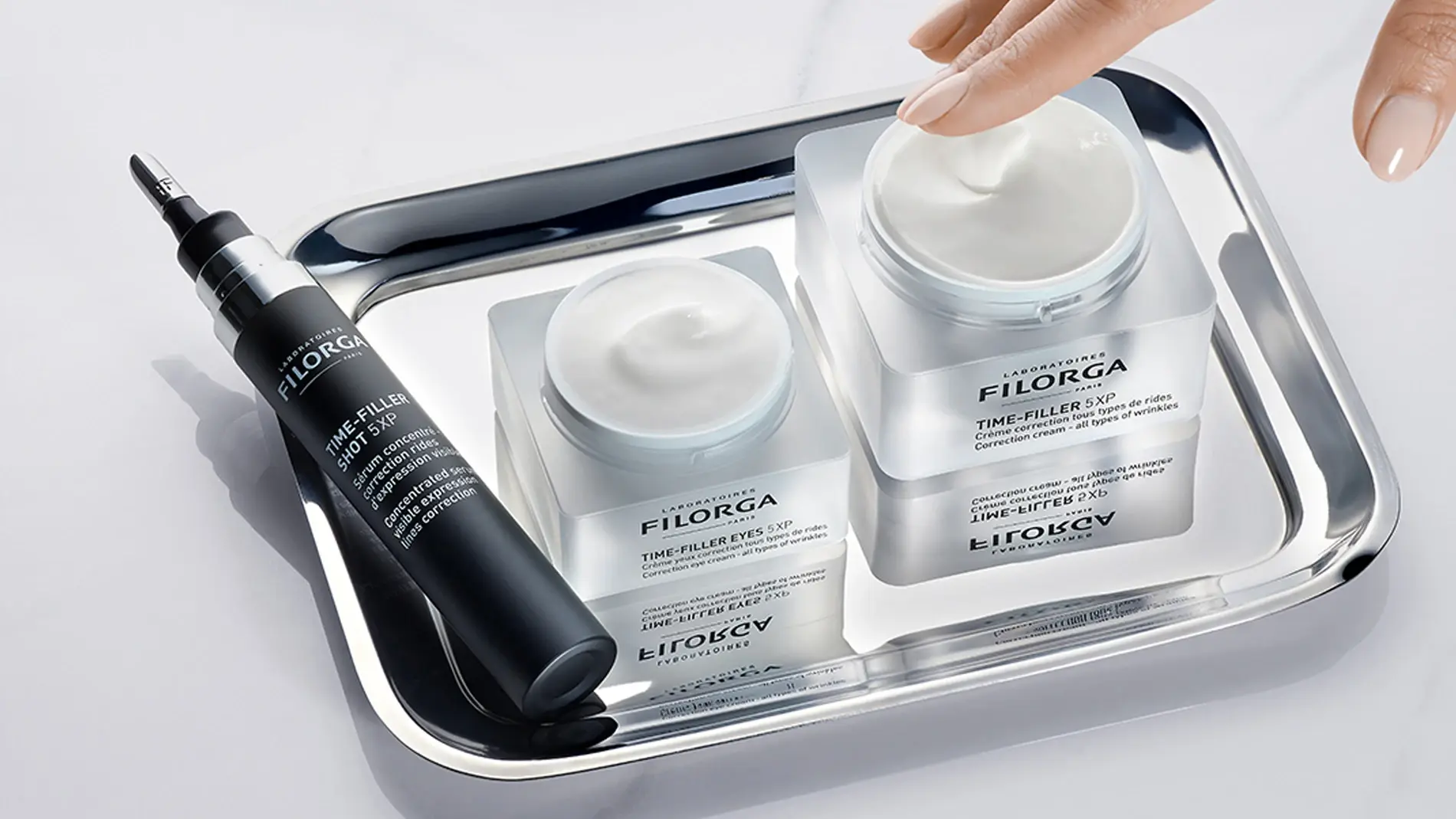 En hånd berører en krukke med Filorga "TIME-FILLER 5XP" krem, som står på et sølvbrett sammen med en mindre krukke og en tube med "TIME-FILLER EYES 5XP".