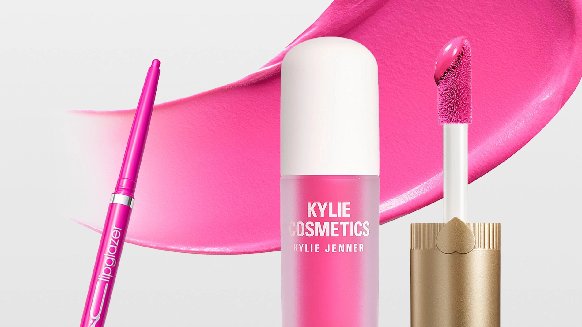 Kylie Cosmetics Supplе Kiss Lip Glaze -huulikiilto yhdessä vaaleanpunaisen "lipglazer"-huultenrajauskynän kanssa, applikaattori ja levitetty kiiltotekstuuri taustalla. Pakkauksessa lukee "KYLIE COSMETICS KYLIE JENNER SUPPLE KISS LIP GLAZE".