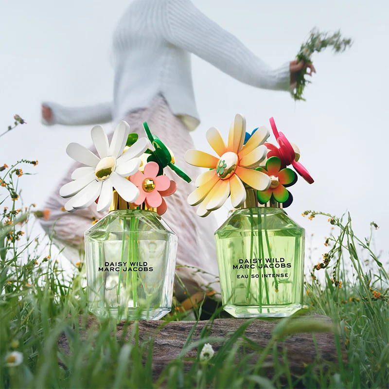 To Daisy Wild Marc Jacobs-parfymeflasker med fargerike blomster som korker. Bak flaskene en person som holder blomster i hånden, kledd i hvit genser og rosa bukser. Bakgrunnen har grønt gress og små blomster.