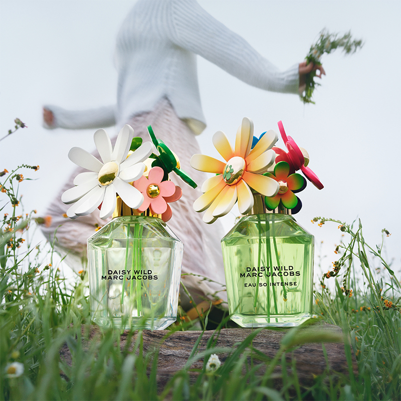Två Daisy Wild Marc Jacobs-parfymflaskor med färgglada blommor som korkar. Bakom flaskorna en person som håller blommor i handen, klädd i vit tröja och rosa byxor. Bakgrunden har grönt gräs och små blommor.