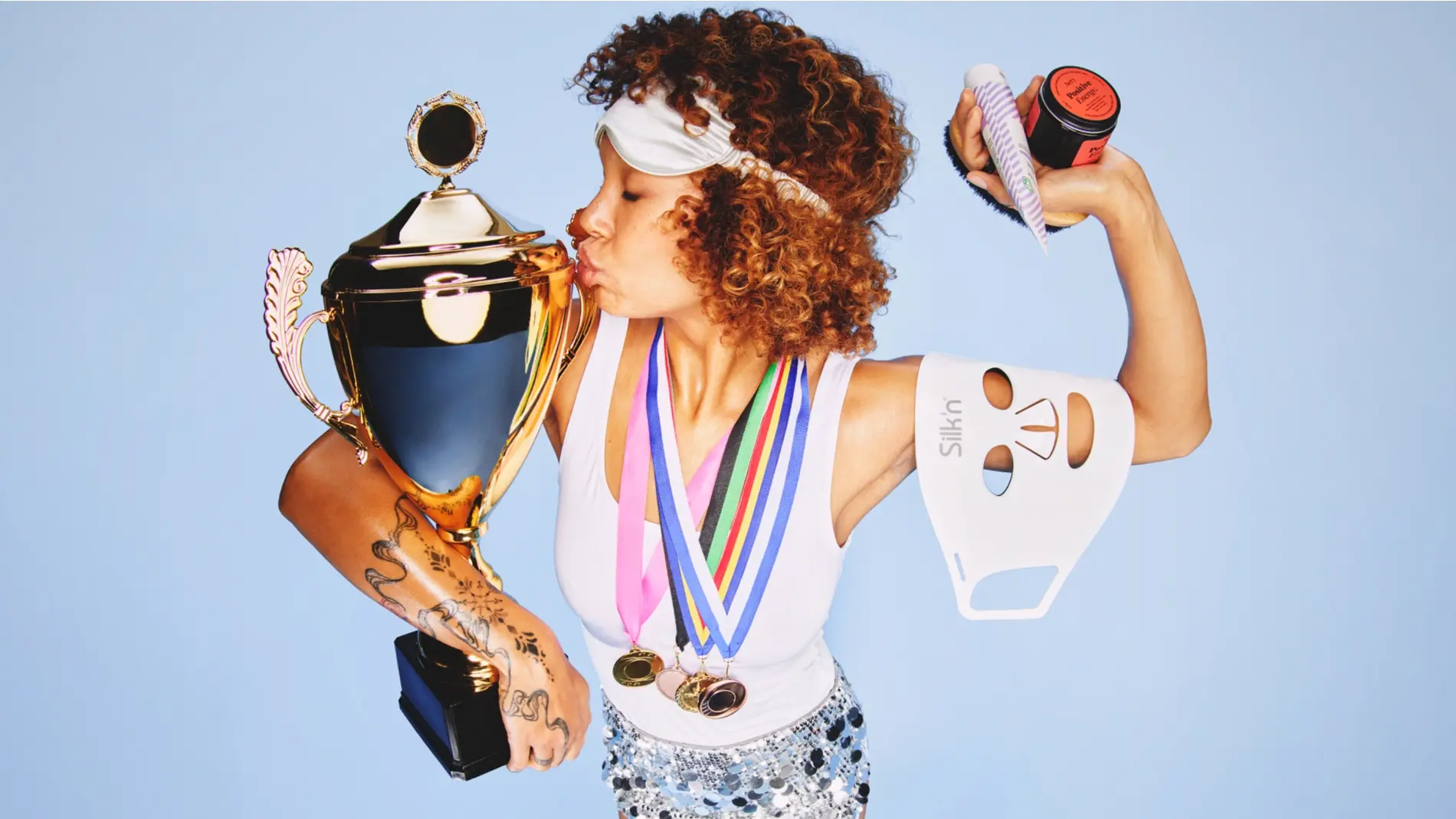 Person med lockigt hår och sovmask kysser en stor guldfärgad pokal och bär flera medaljer runt halsen. Personen håller även en vit Silkn LED-mask och hudvårdsprodukter mot en ljusblå bakgrund, med sportig och glamorös beauty-känsla.