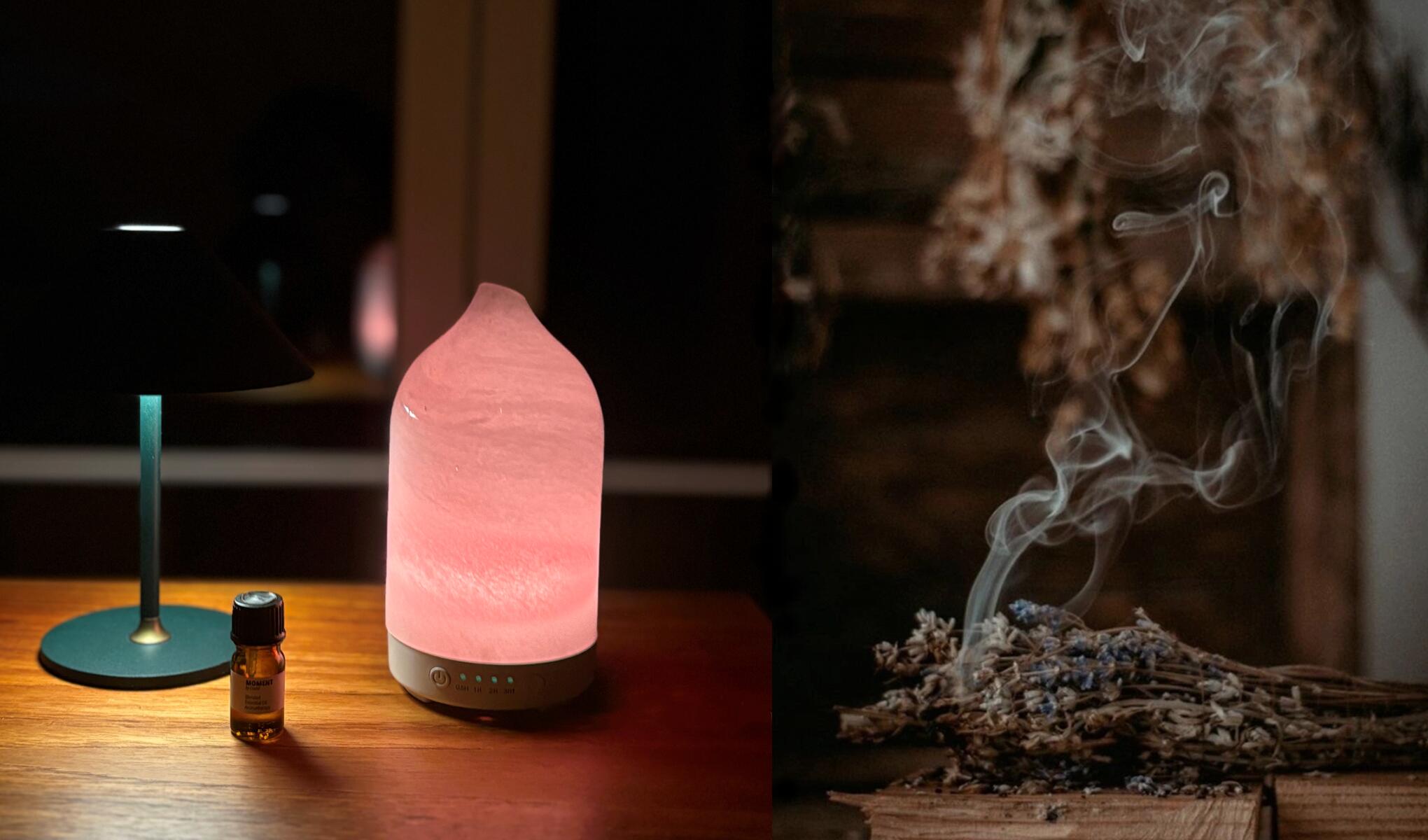 Doftdiffuser med mjukt rosa ljus står på ett träbord bredvid en liten flaska eterisk olja och en bordslampa, med knappar synliga på basen. Till höger syns rykande torkade växter som sprider doft, vilket förstärker en aromaterapi- och hemmaspa-känsla.
