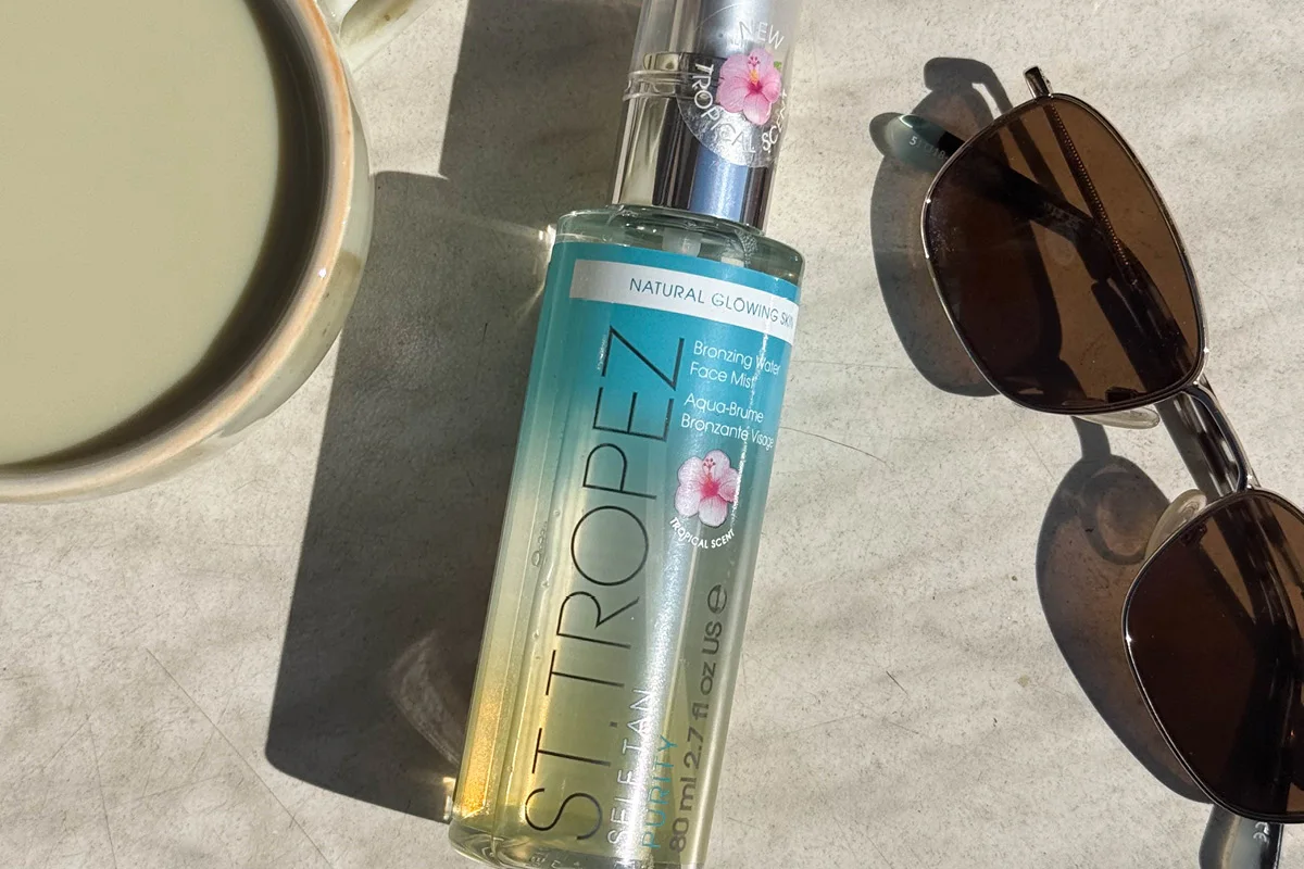 St. Tropez Self Tan Purity Bronzing Water Face Mist pöydällä auringonvalossa aurinkolasien ja kupin vieressä.