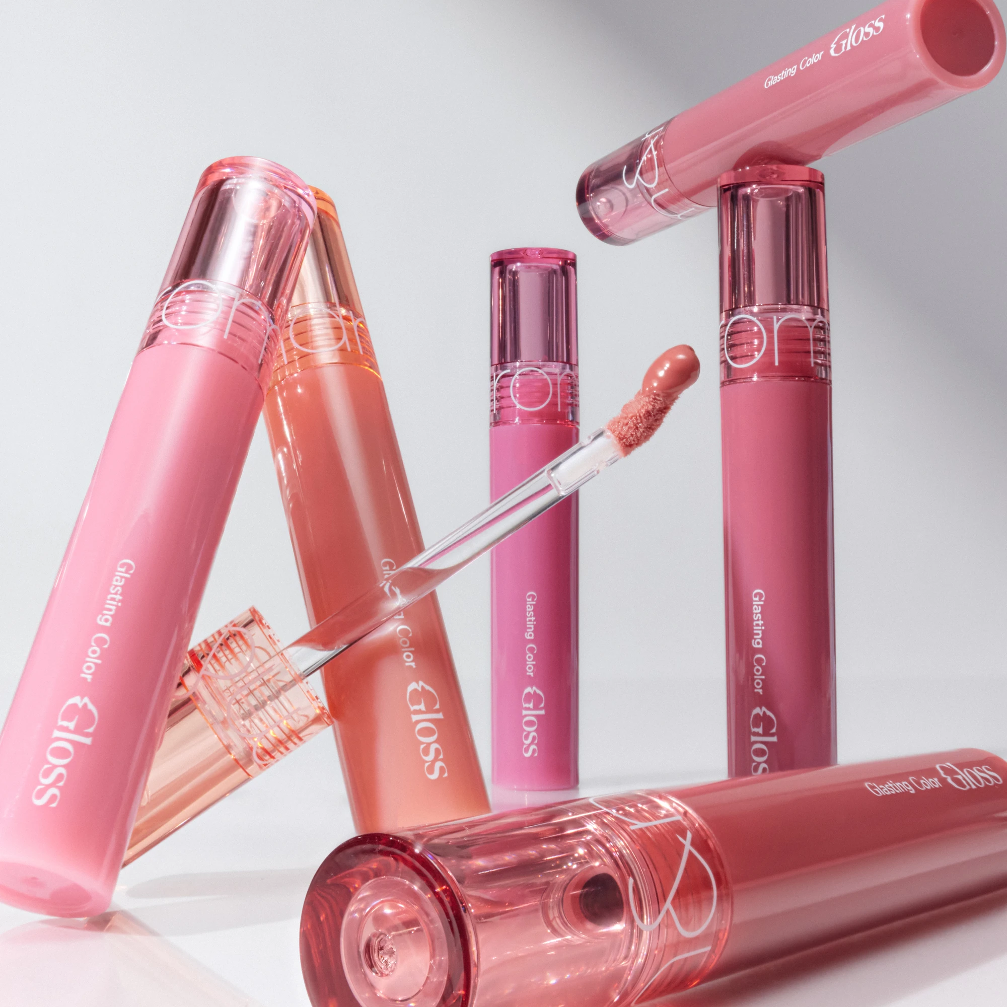 Utvalg av 'Glasting Color Gloss' leppeglosser i rosa og ferskenfarger, én med applikatoren eksponert.