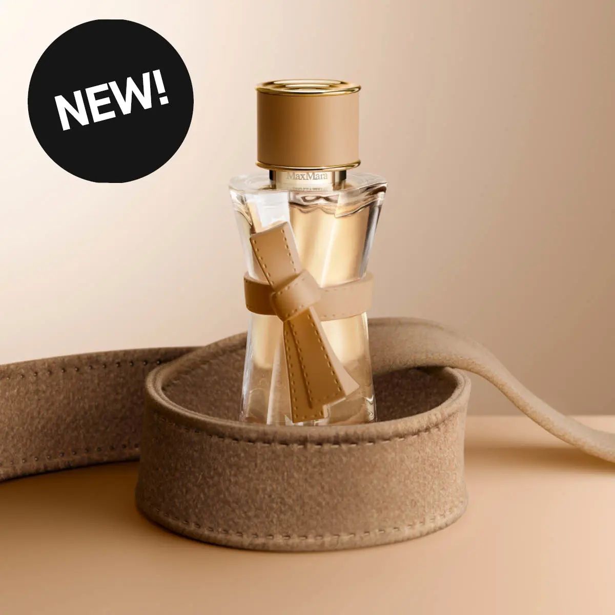Max Mara-parfyme i gjennomsiktig flaske med gullfarget kork og beige sløyfedetalj, plassert i et mykt beige etui. Svart sirkel med den hvite teksten «NEW!».