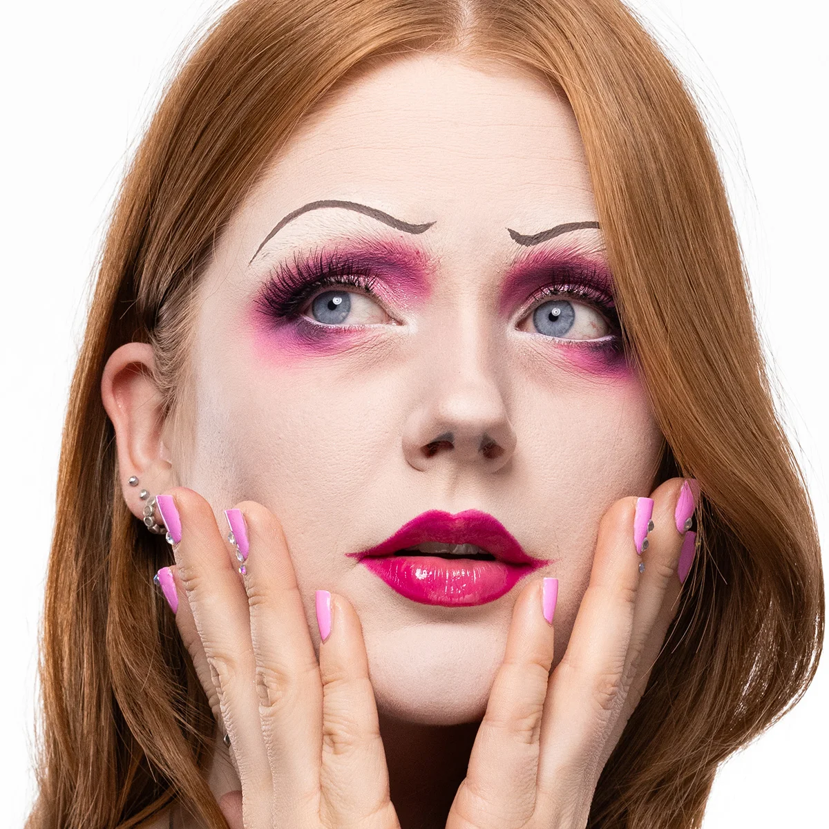 En person med rosa och lila ögonskugga, långa fransar och tunna svartmålade ögonbryn, tillsammans med glansiga ceriserosa läppar och rosa dekorerade naglar som ramar in ansiktet i en dramatisk, Tim Burton-inspirerad look.