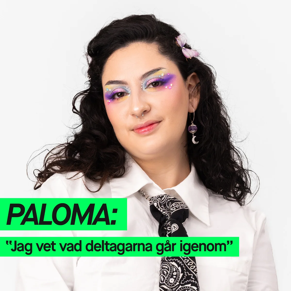 En person med lekfull och färgstark ögonmakeup i lila och pastelltoner samt dekorerande ansiktsdetaljer poserar i vit skjorta och svart mönstrad slips, med texten ”PALOMA: Jag vet vad deltagarna går igenom”.
