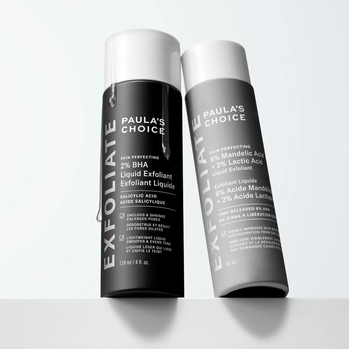 Paula's Choice - To hudpleieprodukter står på en hvit overflate. En svart flaske og en grå flaske, begge med hvite korker.