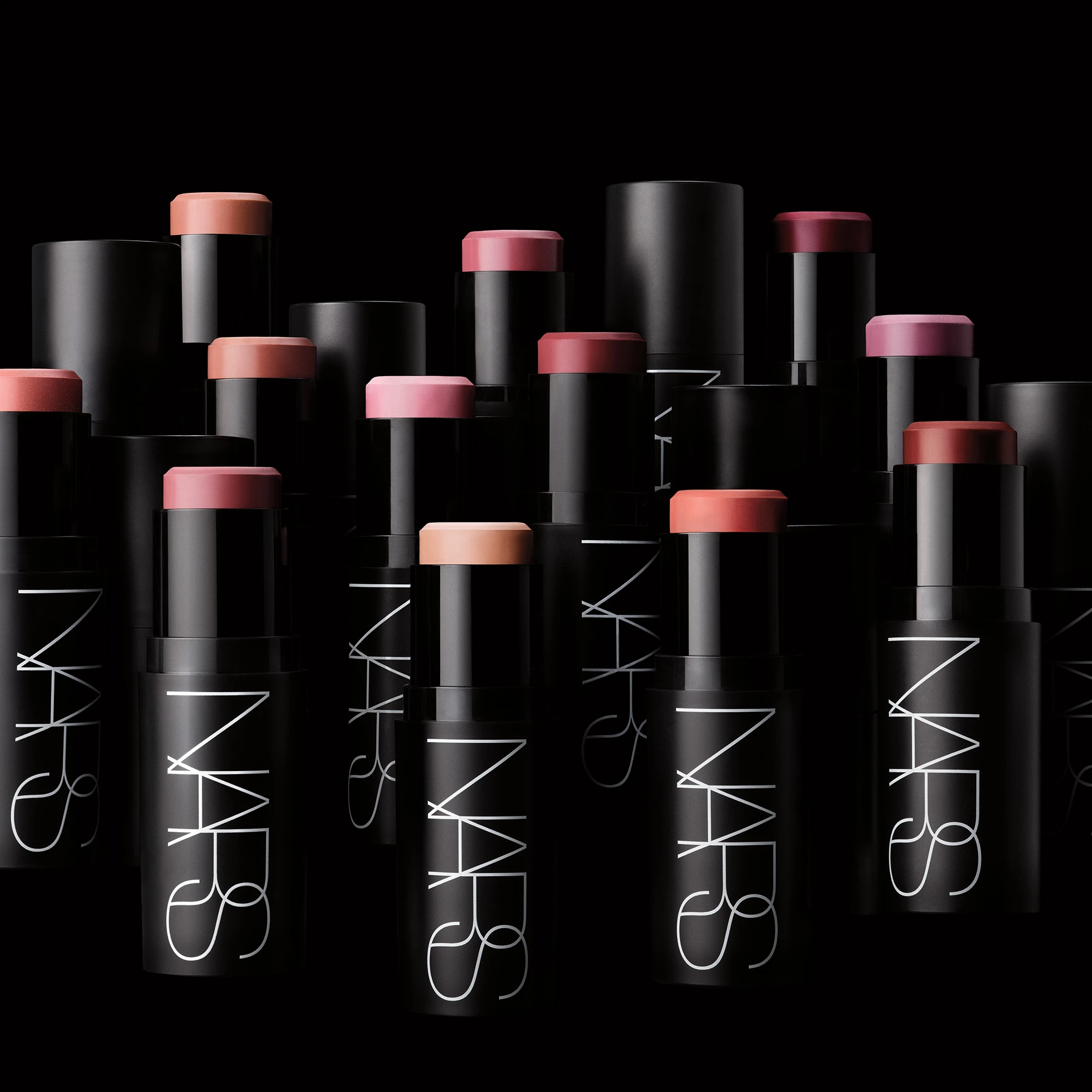 Useita NARS cream blush -puikkoja vaaleanpunaisen, persikan, nude-sävyjen ja marjasävyjen väreissä on aseteltu mustaa taustaa vasten. Mustissa pakkauksissa näkyy valkoinen “NARS”-logo, ja esillä on useita voidemaisia poskipunasävyjä.