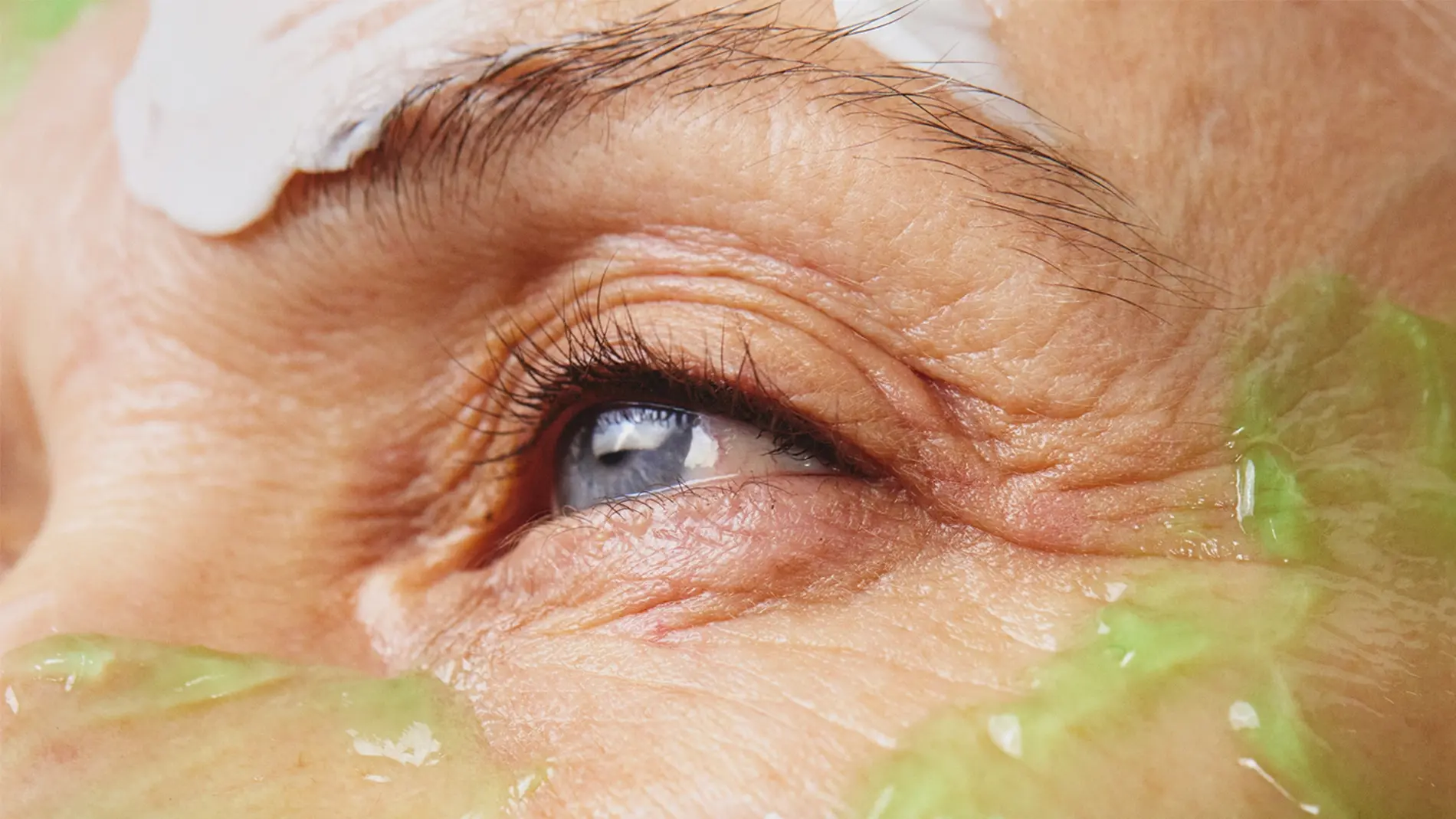 Närbild av ett öga med synliga hudvårdsprodukter applicerade runt ögat och på kinden. En grön gelmask och en vit exfolierande ansiktsmask med svarta korn framhäver hudvård och ansiktsbehandling.