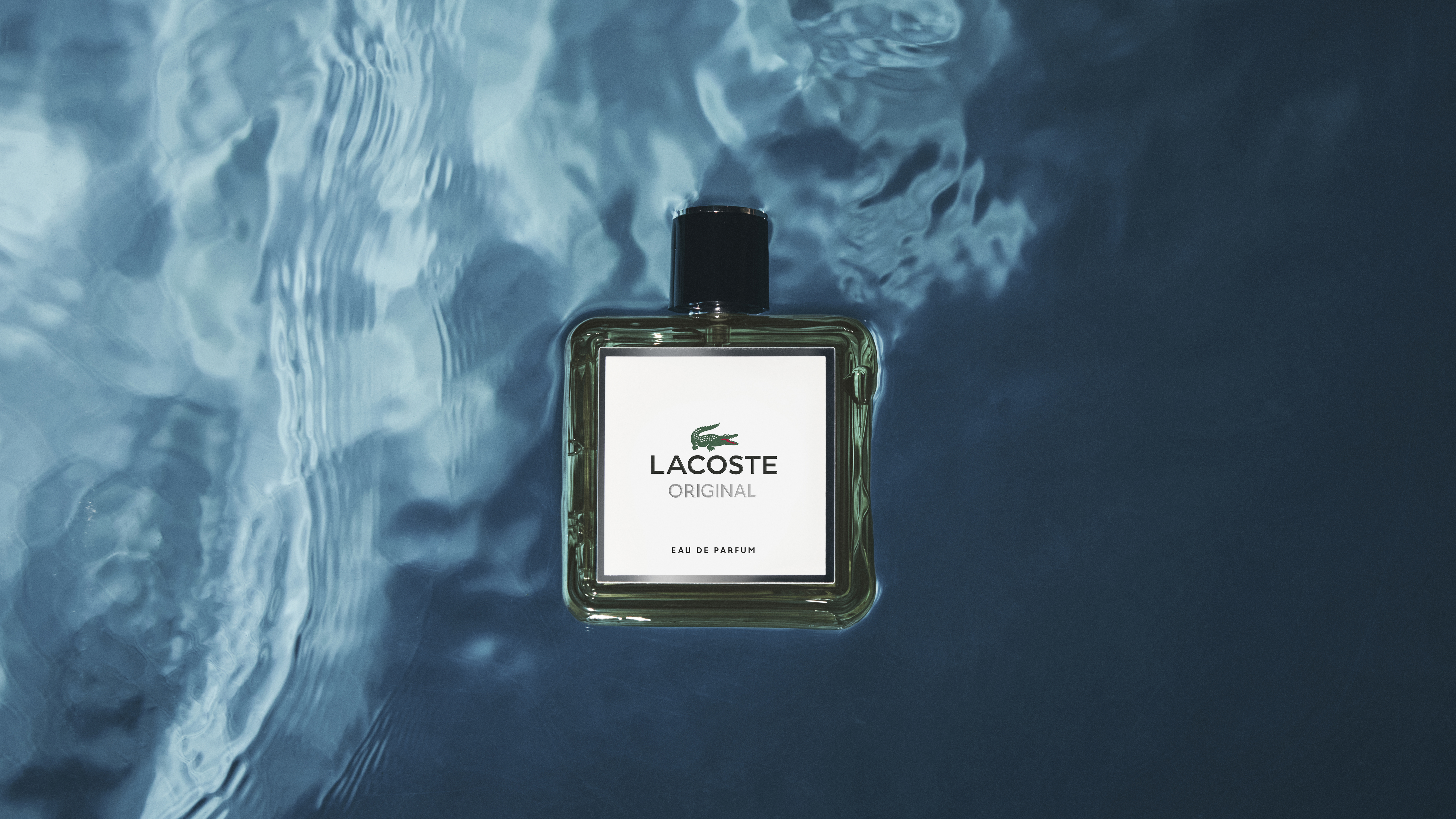 Lacoste Original -hajuvesipullo kelluu veden pinnalla, jossa on sinisiä värejä ja heijastuksia.
