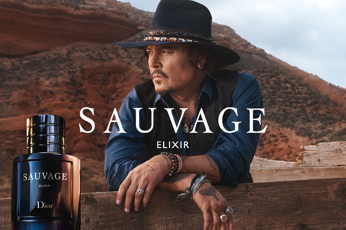 Sauvage Elixir -tuoksu Diorilta. Mies seisoo ulkona, pukeutuneena siniseen paitaan ja mustaan liiviin, sekä mustaan hattuun. Taustalla on karu maisema, jossa näkyy punertavia kallioita. Vasemmassa alakulmassa on Sauvage Elixir -pullo.