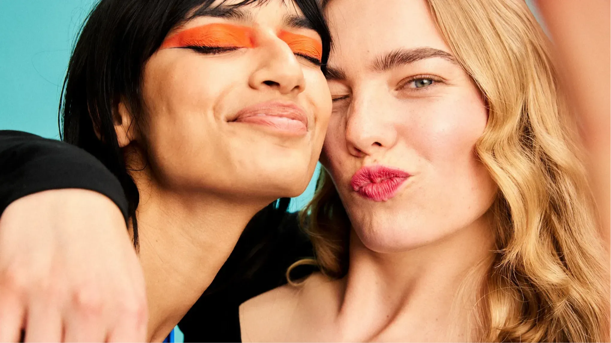 To unge kvinner poserer tett sammen med smil og kyssemunner. Den ene har kraftig oransje øyenskygge og svart hår, mens den andre har bølgete blondt hår og rosa leppestift. Bakgrunnen er lyseblå.