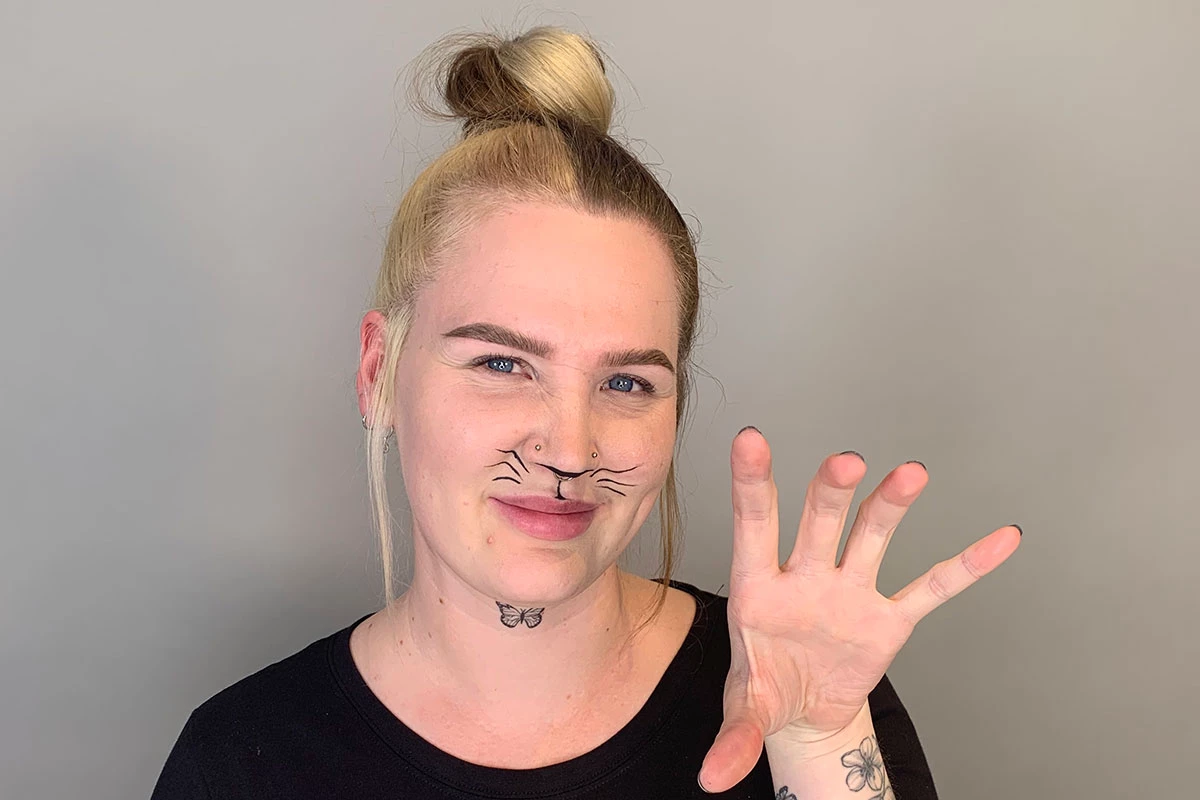 Nainen, jolla on musta nenänpää, viiva nenän alla ja silmänrajauskynällä poskille piirretyt viikset, nostaa kättään kuin kissa.