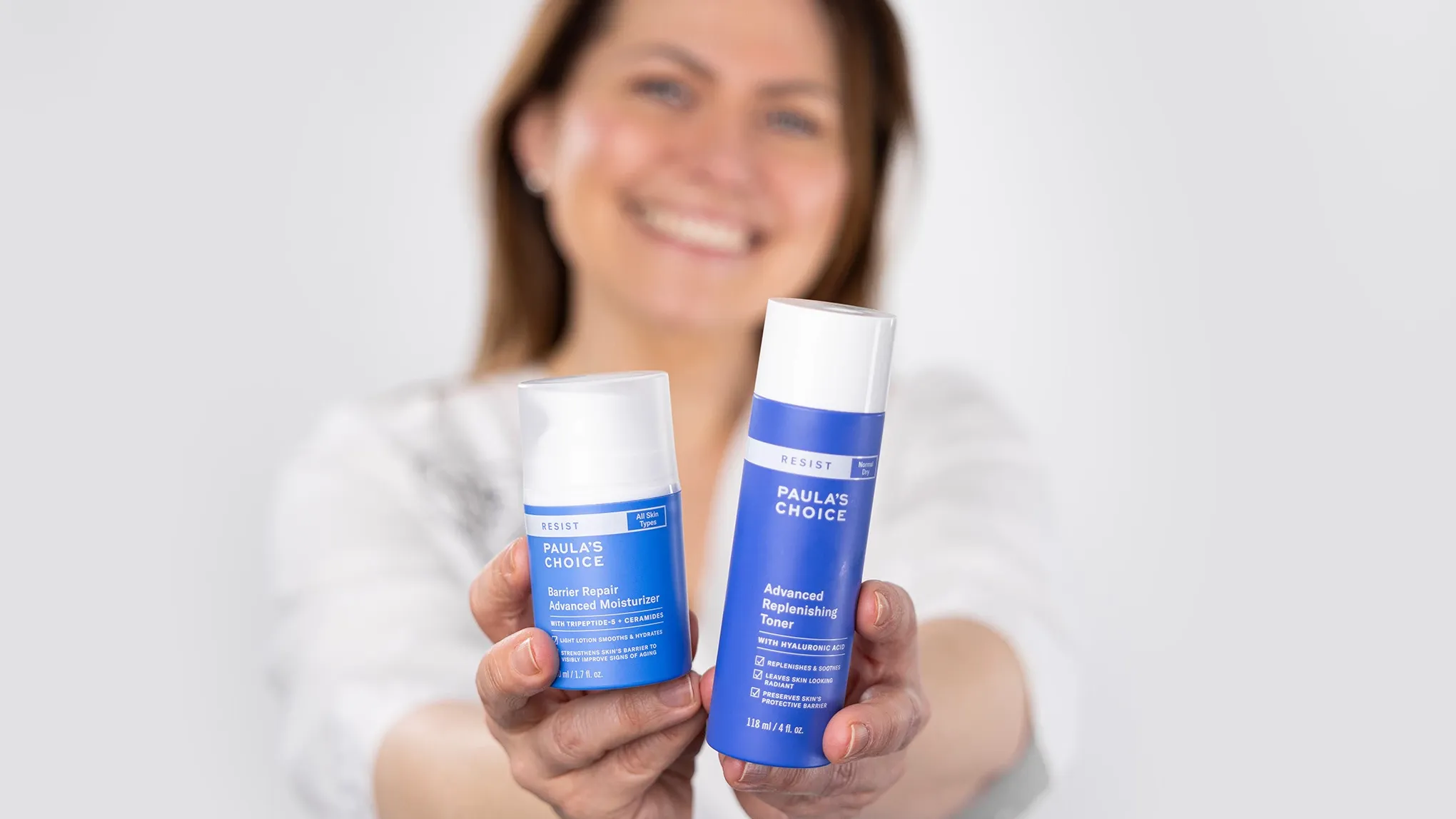 To blå produkter fra Paula's Choice foran en uklar kvinne med et stort smil mot en hvit bakgrunn.