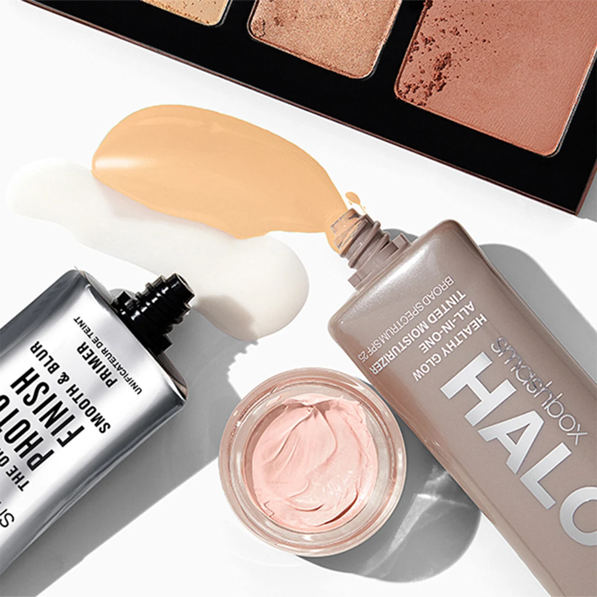 En samling sminkprodukter från Smashbox placerade på en vit yta. En öppen palett syns i övre delen av bilden. En silverfärgad tub och en beige tub har kräm som har runnit ut bredvid dem. En liten burk med rosa kräm är placerad i mitten. Produkterna har förpackningar i silver, beige och rosa.
