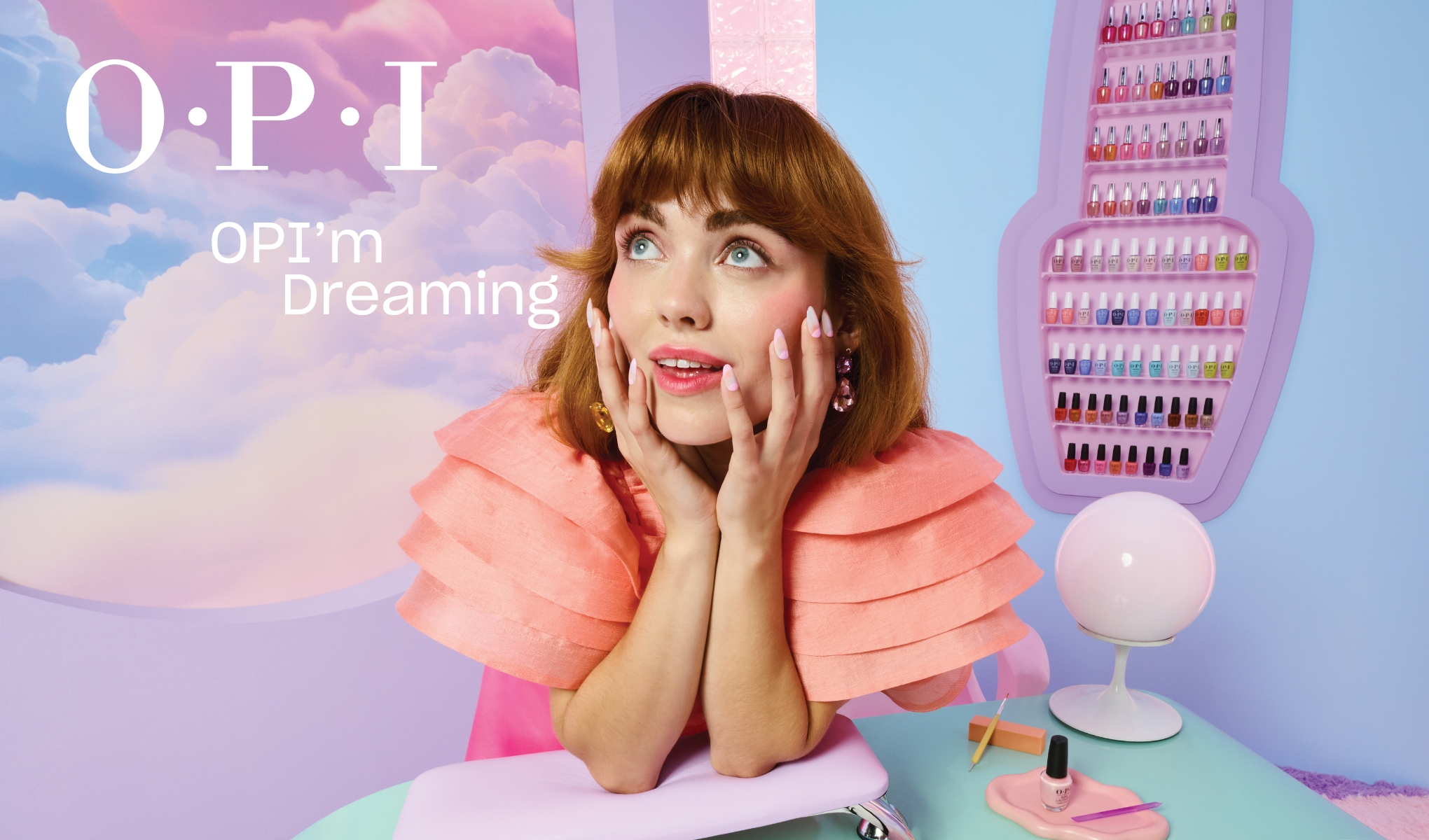 Person sittande vid ett bord med nagellacksprodukter från OPI. Bakgrunden är färgglad med moln och en hylla fylld med olika färger av nagellack. Texten "OPI OPI'm Dreaming" syns till vänster.