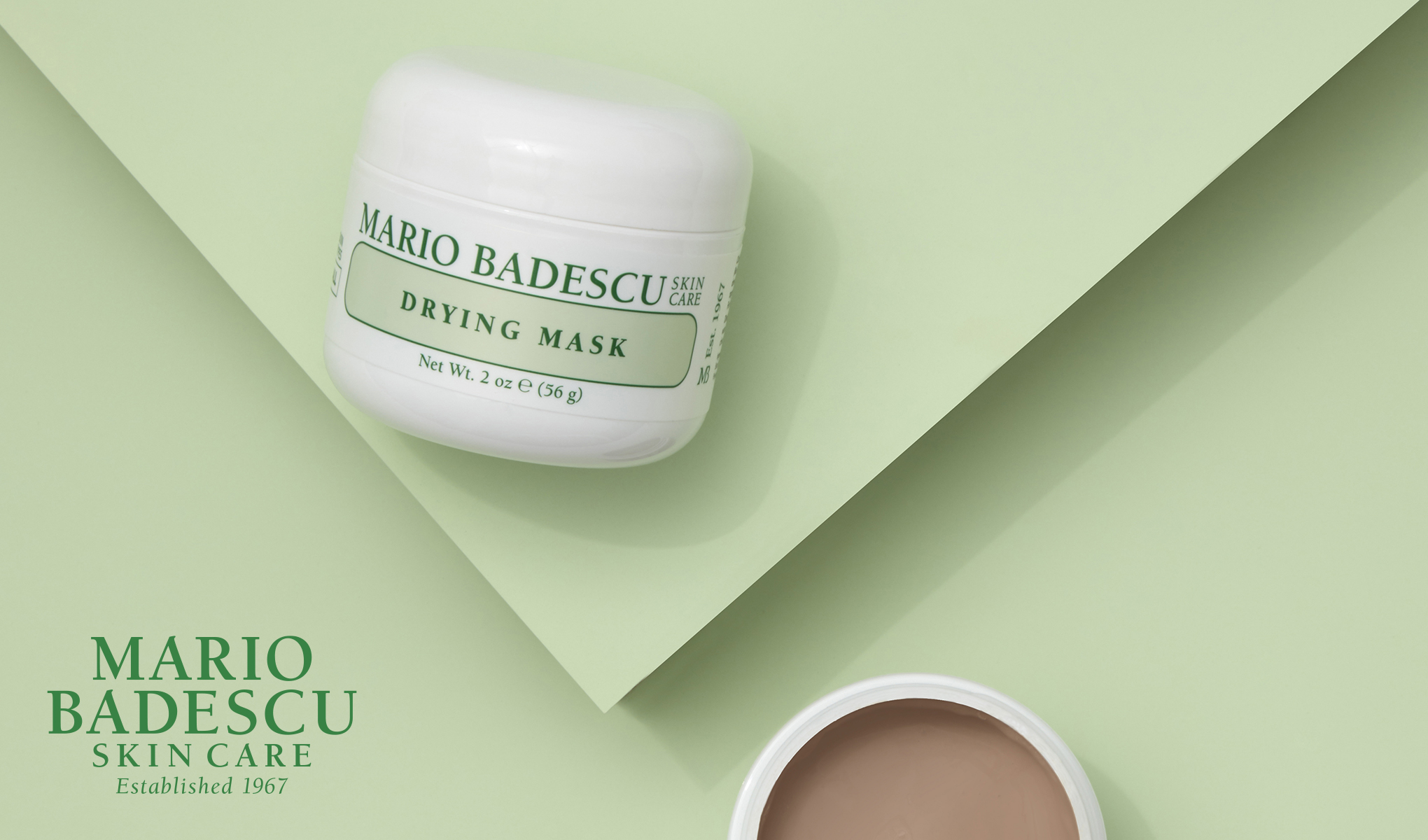 Valkoinen purkki, jossa on Mario Badescun kuivausnaamio vaaleanvihreällä pohjalla. Purkki on merkitty vihreillä kirjaimilla. Vasemmassa alakulmassa on teksti ”Mario Badescu Skin Care Established 1967”. Avoin purkki, jossa on naamio, näkyy myös kuvan oikeassa alakulmassa.