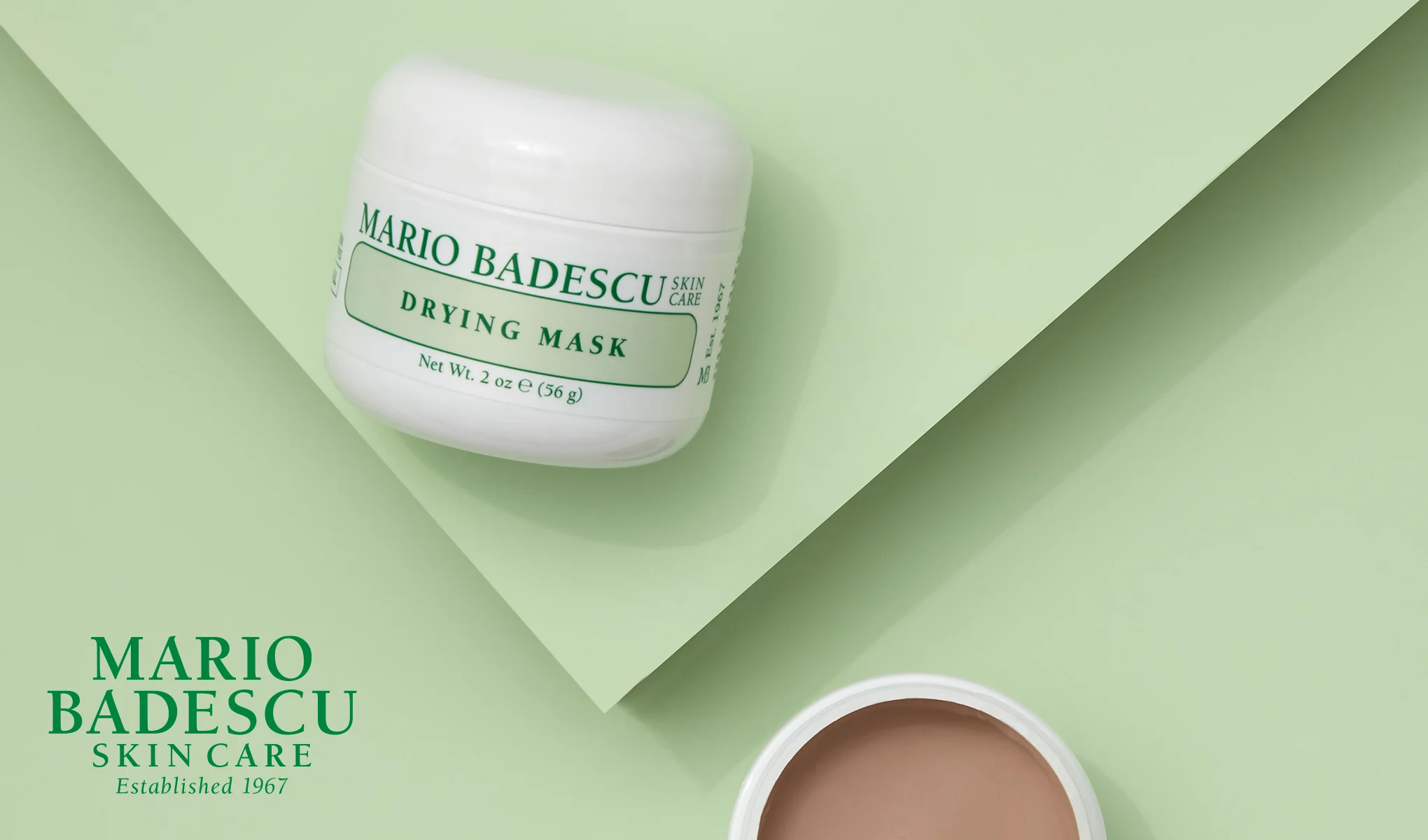 En vit burk med Mario Badescus Drying Mask placerad på en ljusgrön bakgrund. Burken är märkt med gröna bokstäver. I nedre vänstra hörnet finns texten "Mario Badescu Skin Care Established 1967". En öppen burk med masken syns också i bildens nedre högra hörn.