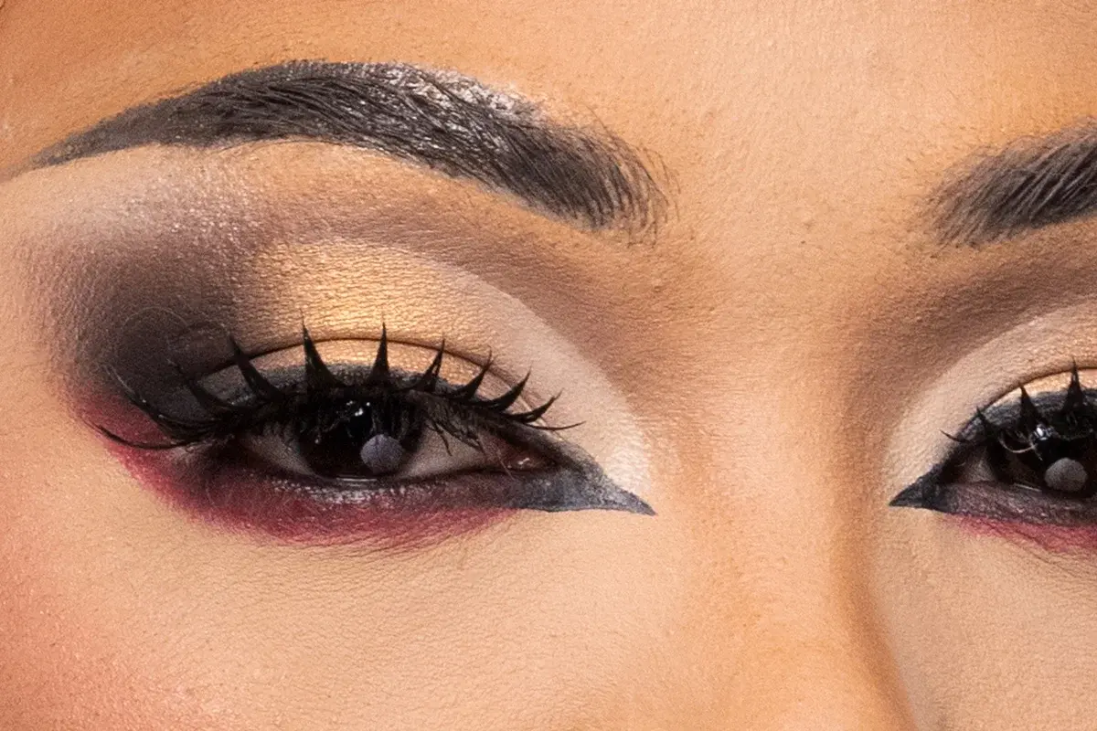 Närbild av ögonmakeup med dramatisk kontrast mellan guld, svart och vinrött, skarpa eyelinerdetaljer och långa fransar som framhäver en uttrycksfull look.