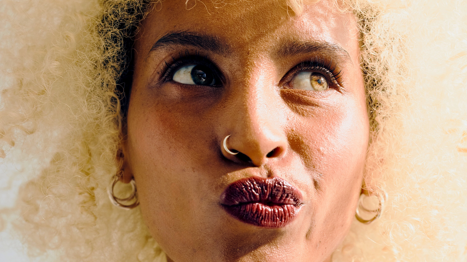 Nærbilde av en person med krøllete hår som ser opp til siden. Personen har markerte øyenbryn, mascara, mørk leppestift, nesering og øredobber mot en lys bakgrunn.