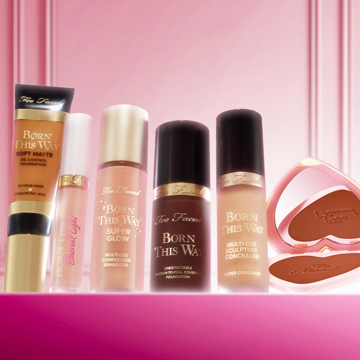 Too Faced-sminkeprodukter på rekke foran rosa bakgrunn, inkludert foundation, concealer og bronzer.