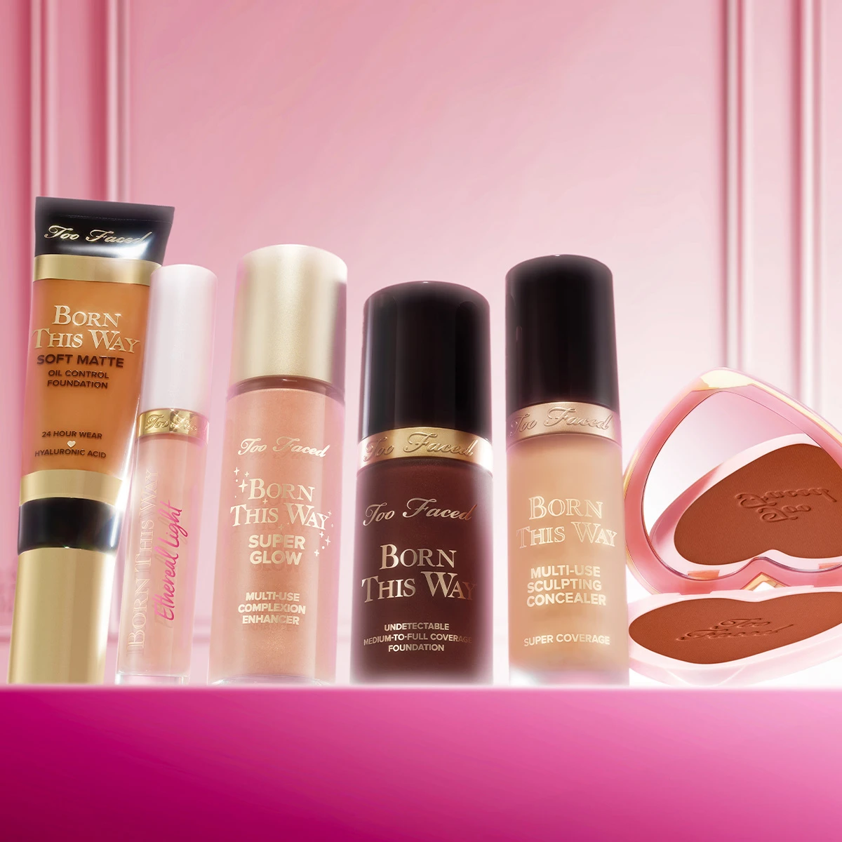 Too Faced -kasvomeikkejä rivissä vaaleanpunaisella taustalla, mukana meikkivoide, peitevoide ja aurinkopuuteri.