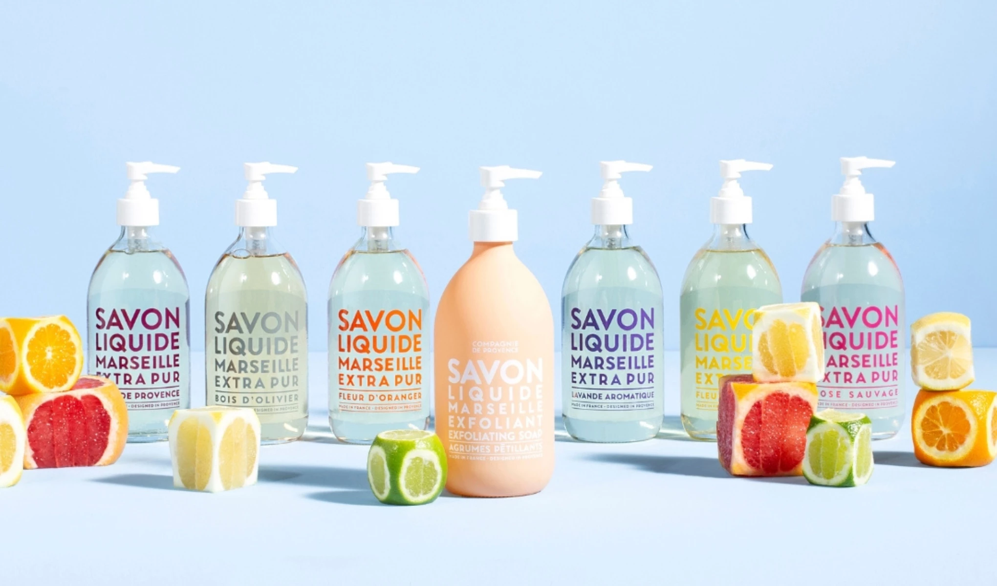 Sju produkter fra Compagnie de Provence i forskjellige farger, hvor den i midten er trukket frem, omgitt av delt frukt, mot en blå bakgrunn.
