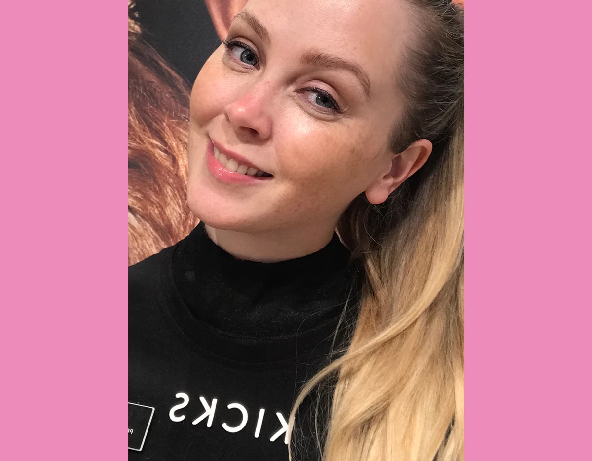 Kvinne i svart t-skjorte med blondt hår som smiler inn i kameraet, med en rosa ramme.