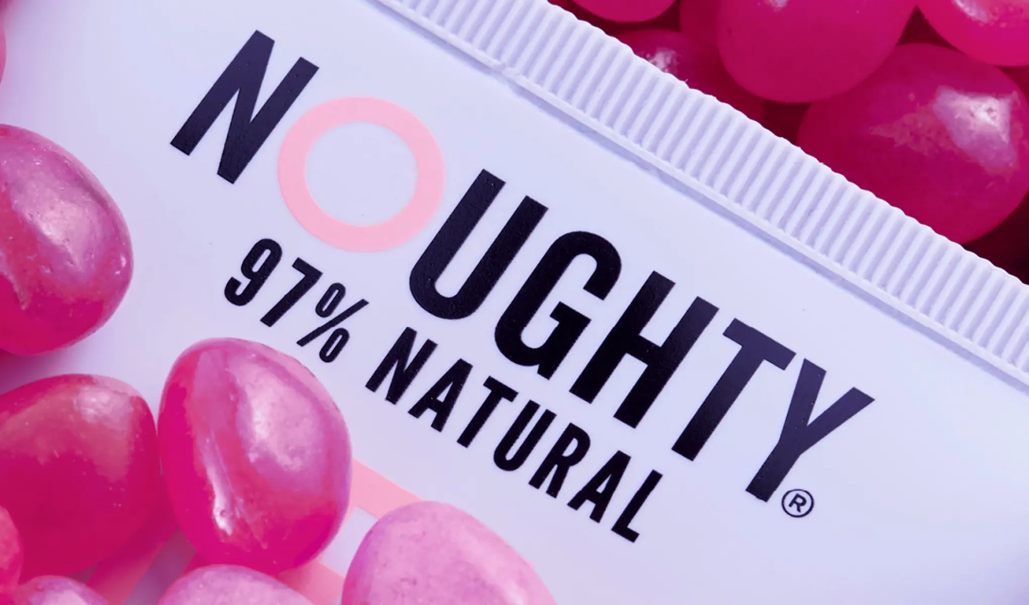 Närbild av en Noughty-produkt med märkningen "97% Natural", omgiven av glansiga rosa stenar för en lekfull och färgstark look.