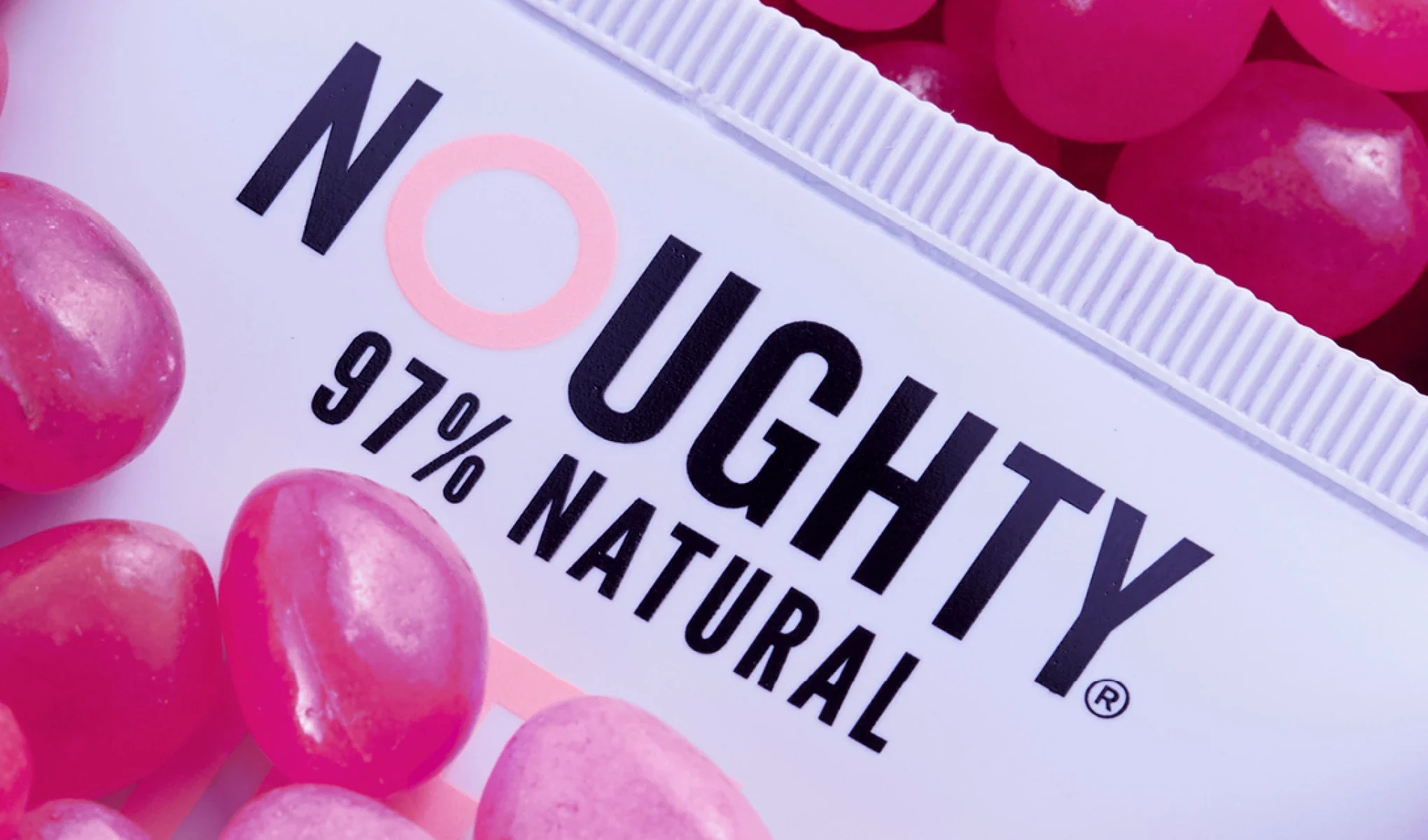 Lähikuva Noughty-tuotteesta, jossa lukee "97% Natural", ympäröitynä kiiltävillä vaaleanpunaisilla kivillä, mikä luo leikkisän ja värikkään ilmeen.
