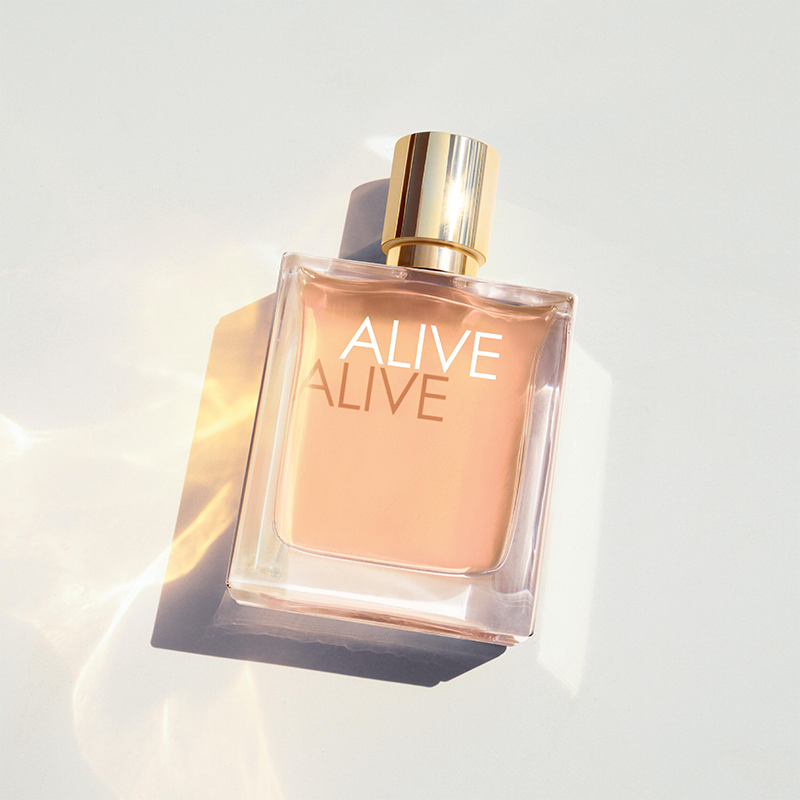 BOSS Alive Eau de Parfum i en gjennomsiktig glassflaske med gullfarget kork ligger mot en lys bakgrunn med lysrefleksjoner. Synlig tekst: "ALIVE" og "ALIVE" på flasken.
