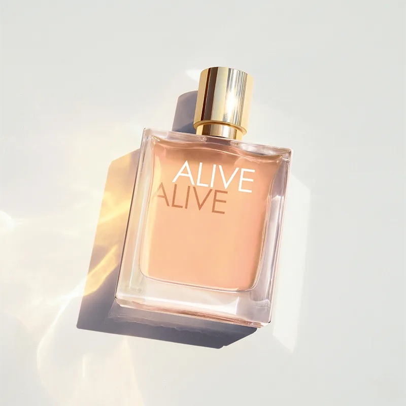 BOSS Alive Eau de Parfum i en gjennomsiktig glassflaske med gullfarget kork ligger mot en lys bakgrunn med lysrefleksjoner. Synlig tekst: "ALIVE" og "ALIVE" på flasken.