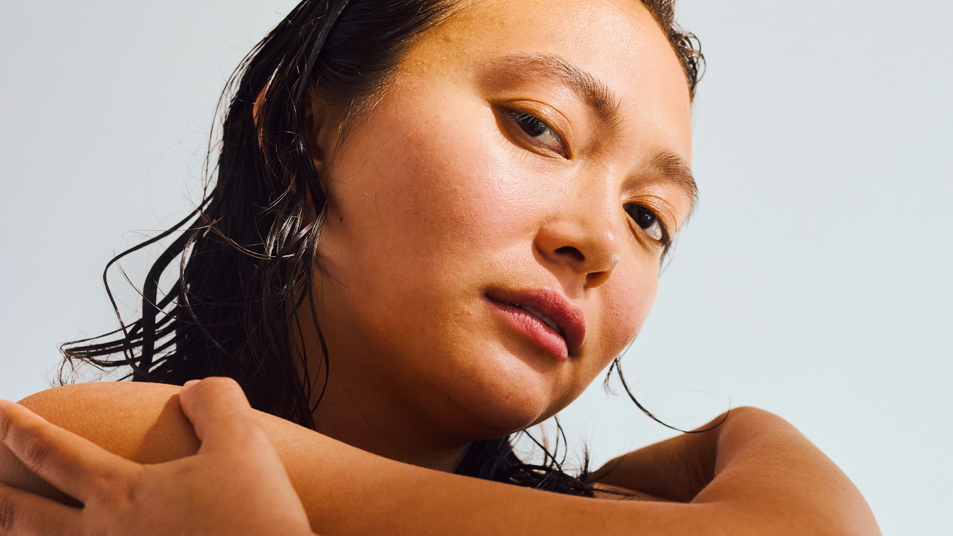 Nærbilde av person med fuktig, bakoverkjemmet hår som krysser armene over skuldrene. Nøytral bakgrunn, naturlig hud og mykt lys.