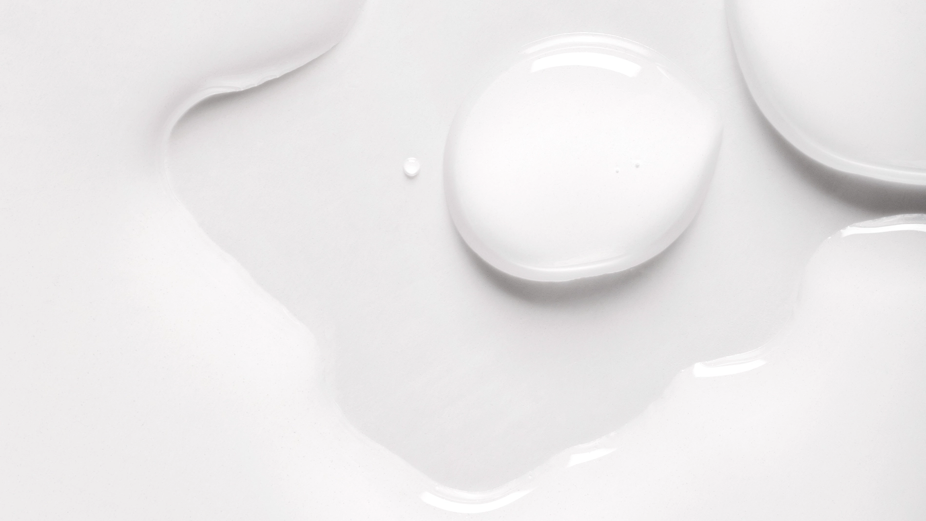 Nærbilde av transparent hudpleieprodukt i flytende form som ligger i myke dråper og tynne lag mot hvit bakgrunn.