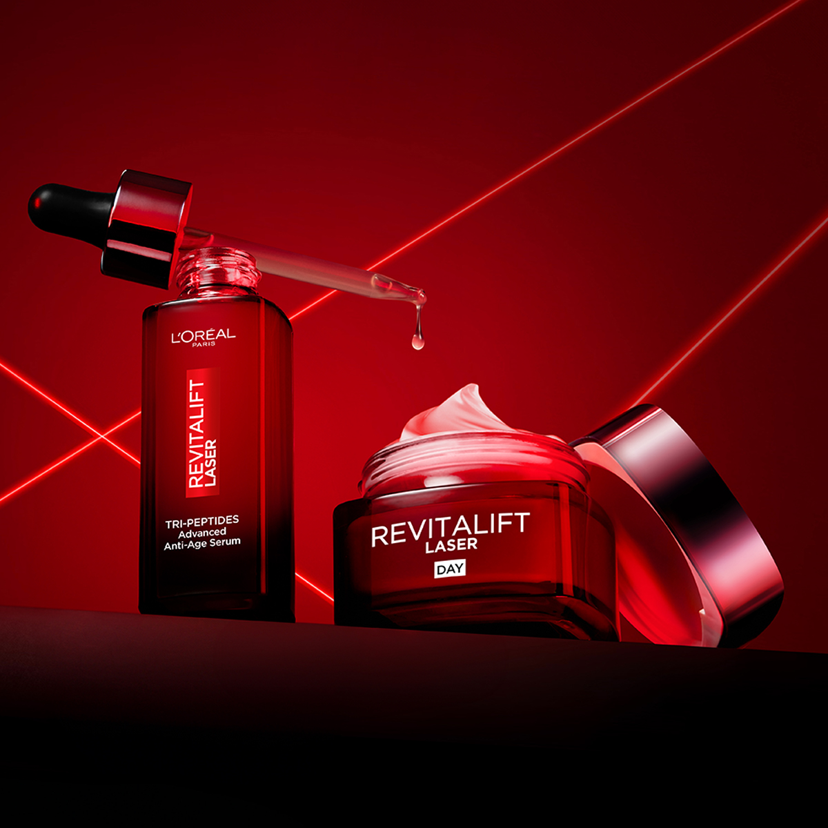 Två Revitalift Laser hudvårdsprodukter från L'Oréal mot en röd bakgrund. Ett serum med pipett och en öppen dagkräm med synlig kräm i locket.
