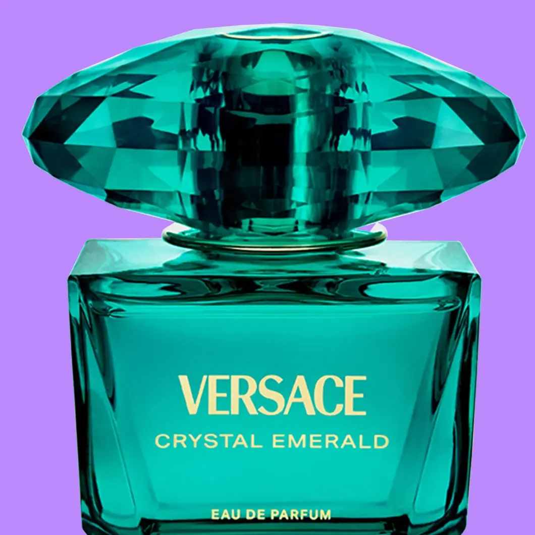 Nærbilde av Versace Crystal Emerald Eau de Parfum i en smaragdgrønn glassflaske med stor fasettert grønn kork og gullfarget tekst «VERSACE CRYSTAL EMERALD EAU DE PARFUM», mot lilla bakgrunn.