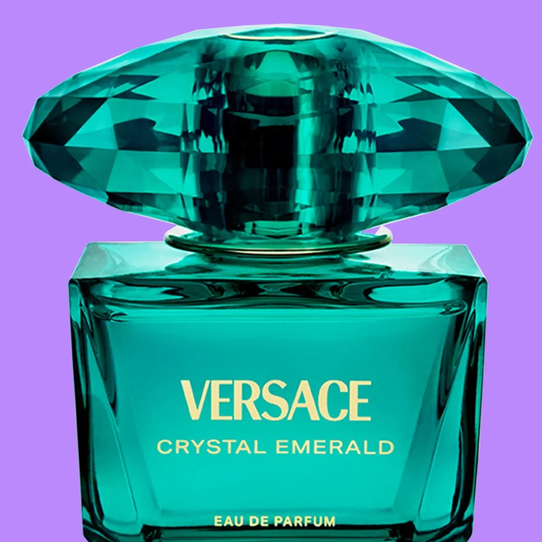Nærbilde av Versace Crystal Emerald Eau de Parfum i en smaragdgrønn glassflaske med stor fasettert grønn kork og gullfarget tekst «VERSACE CRYSTAL EMERALD EAU DE PARFUM», mot lilla bakgrunn.