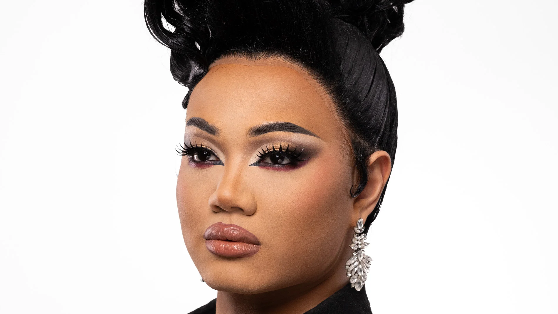 En person med en elegant makeuplook som kombinerar markerad ögonmakeup i guld, svart och vinrött med långa fransar, skulpterad bas och neutrala läppar, kompletterat med stora glittrande örhängen och en uppsatt frisyr.