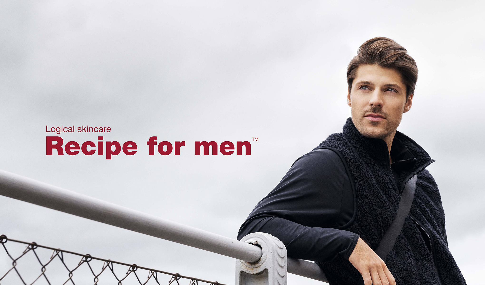Mies nojaa aitaa vasten, yllään musta fleece-liivi ja pitkähihainen paita. Tausta on harmaa ja pilvinen. Kuvan vasemmalla puolella on punaisella teksti ”Logical skincare Recipe for men™”.
