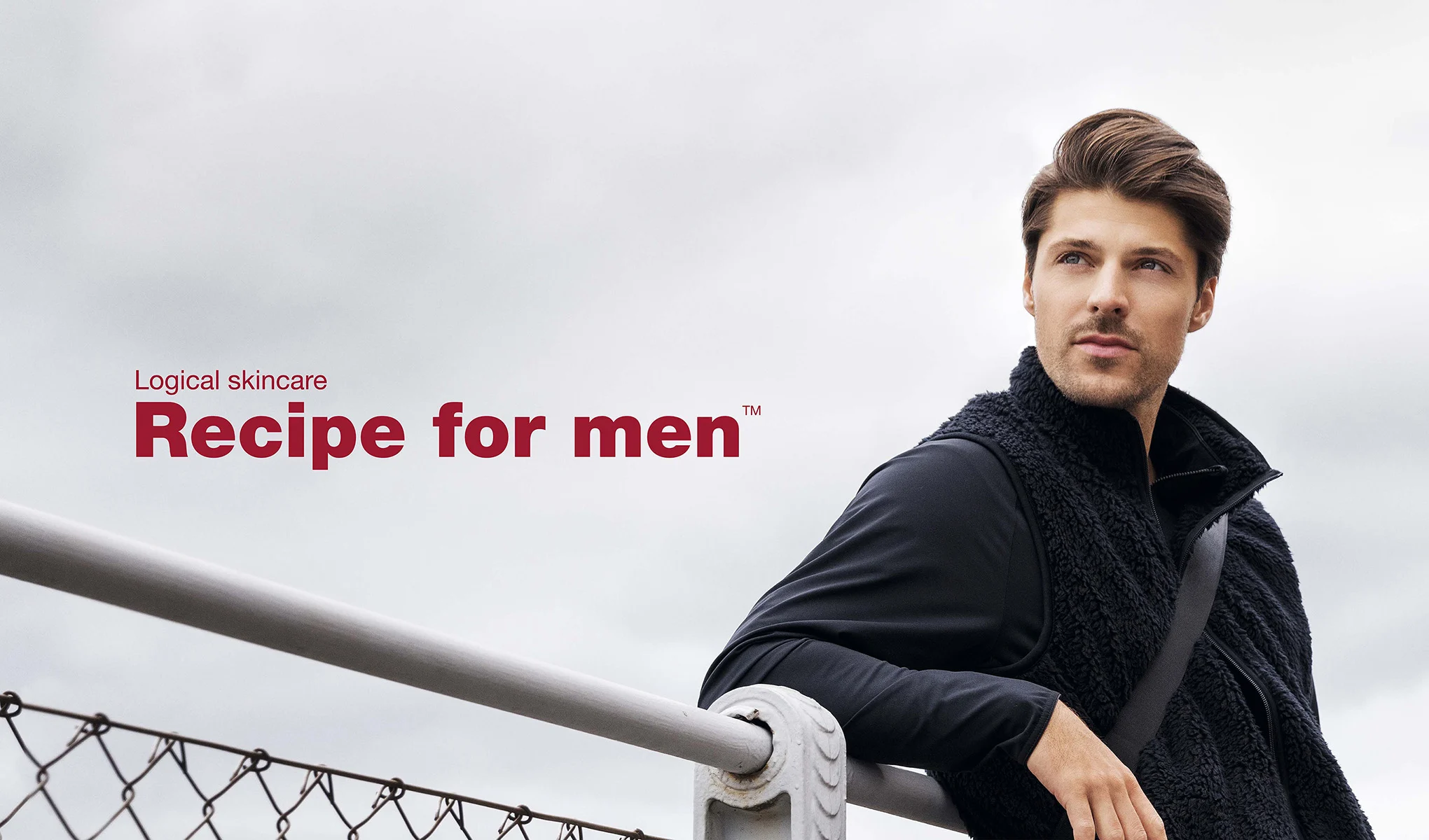 En man står lutad mot ett staket och har på sig en svart fleeceväst och långärmad tröja. Bakgrunden är grå och molnig. Till vänster i bilden finns texten "Logical skincare Recipe for men™" i röd färg.