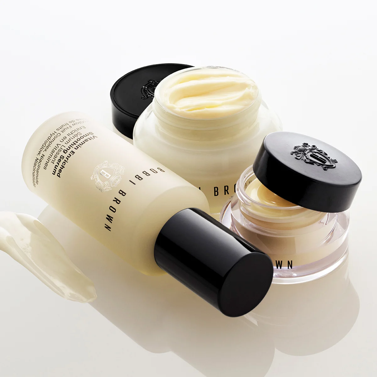 Bobbi Brown Vitamin Enriched Face Base, Vitamin Enriched Smoothing Serum och Vitamin Enriched Eye Base visas tillsammans mot en ljus bakgrund. En öppen ansiktskräm, en öppen ögonkräm och ett serum med svart pumpflaska syns, tillsammans med en krämstrykning. Synlig text: "BOBBI BROWN", "Vitamin Enriched Smoothing Serum", "Face Oil + Vitamin C Complex + Vitamin E", "96% of Fruits" och "Vitamin Enriched Eye Base".