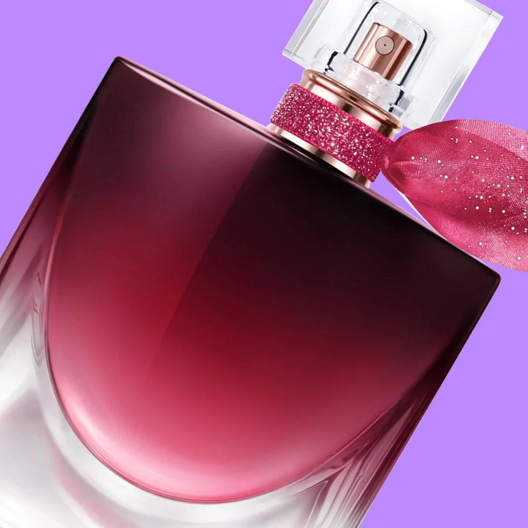 Närbild av en mörkröd parfymflaska med transparent kork, roséguldfärgad spray och glittrande rosa band med rosett mot lila bakgrund.