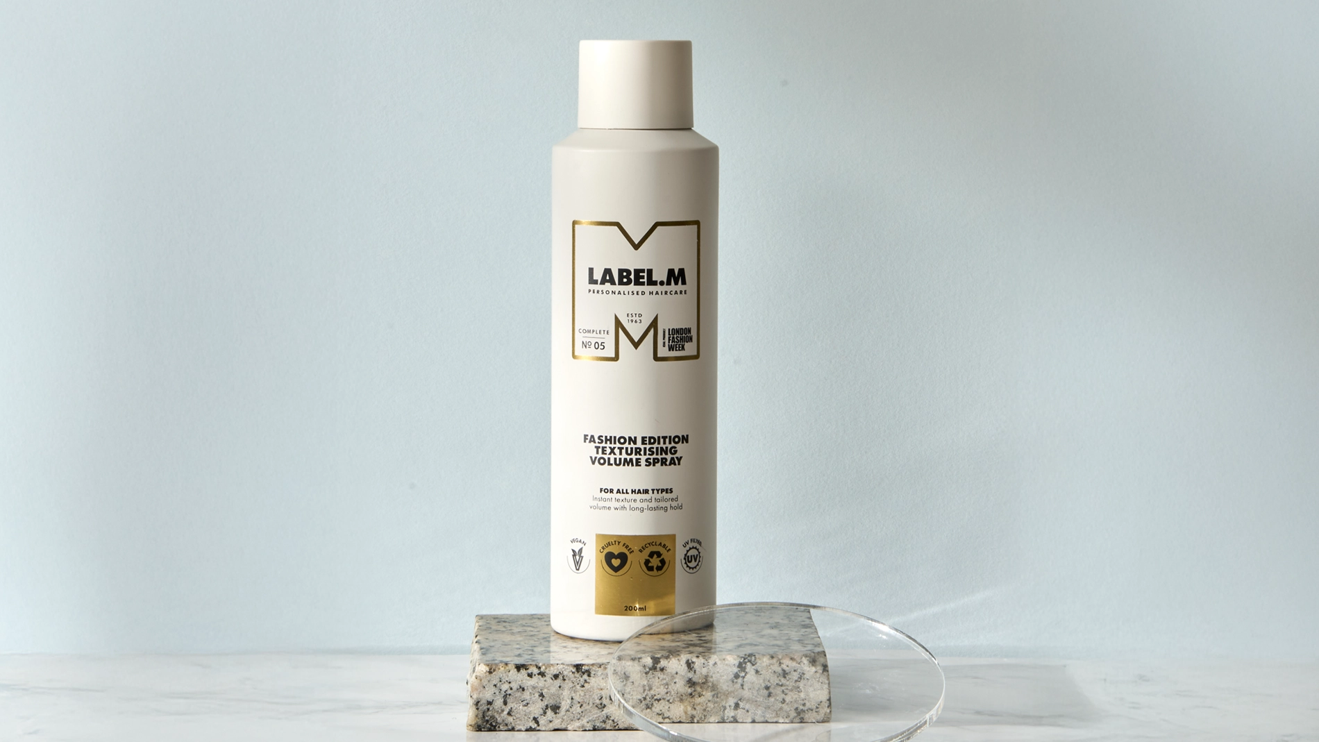 LABEL.M Fashion Edition Texturising Volume Spray i vit flaska placerad på ett stenblock med en transparent akrylskiva framför. Förpackningen visar texten “LABEL.M Personalised Haircare”, “Fashion Edition Texturising Volume Spray”, “For All Hair Types” och volymen 200 ml.