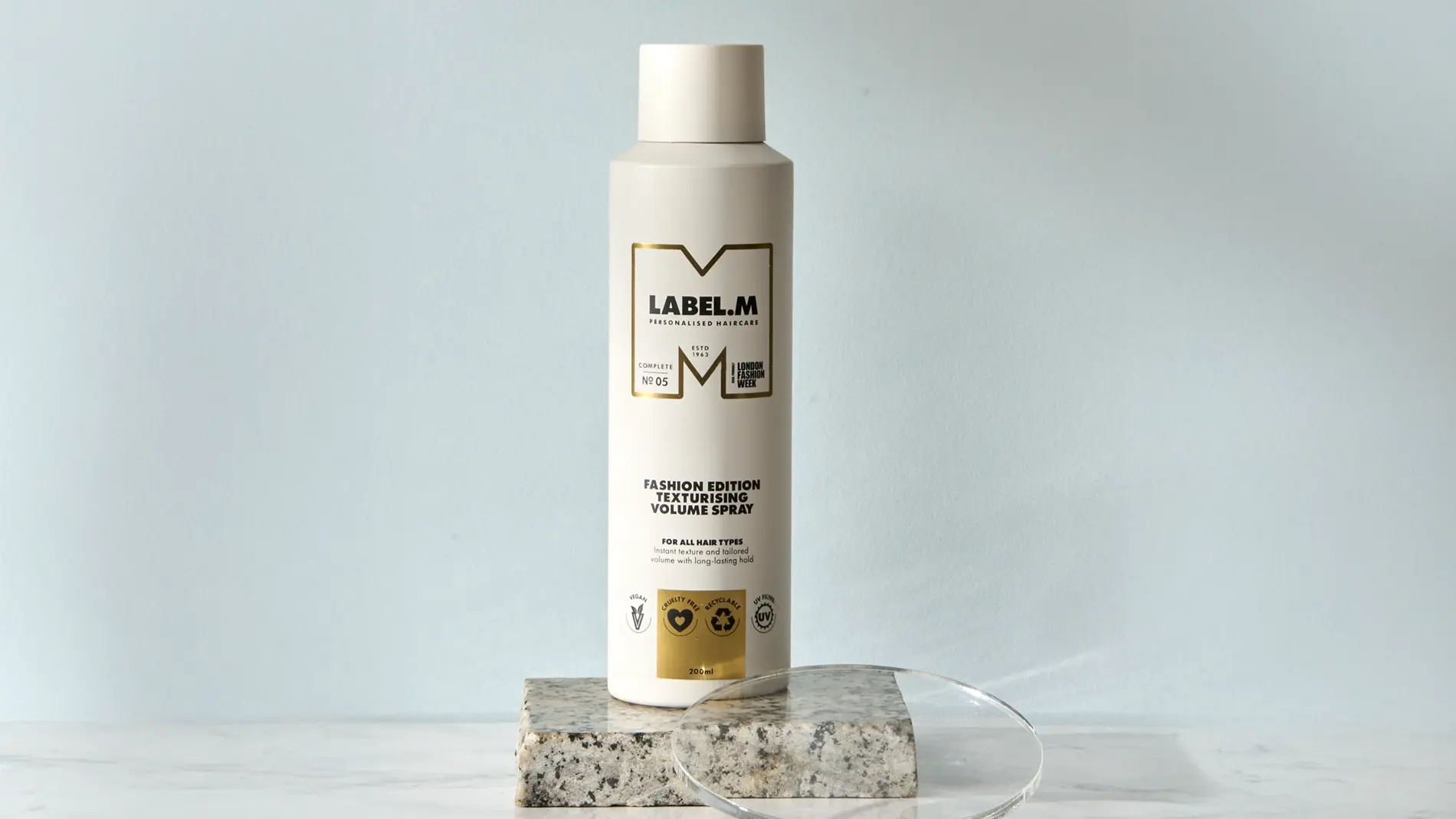LABEL.M Fashion Edition Texturising Volume Spray i vit flaska placerad på ett stenblock med en transparent akrylskiva framför. Förpackningen visar texten “LABEL.M Personalised Haircare”, “Fashion Edition Texturising Volume Spray”, “For All Hair Types” och volymen 200 ml.