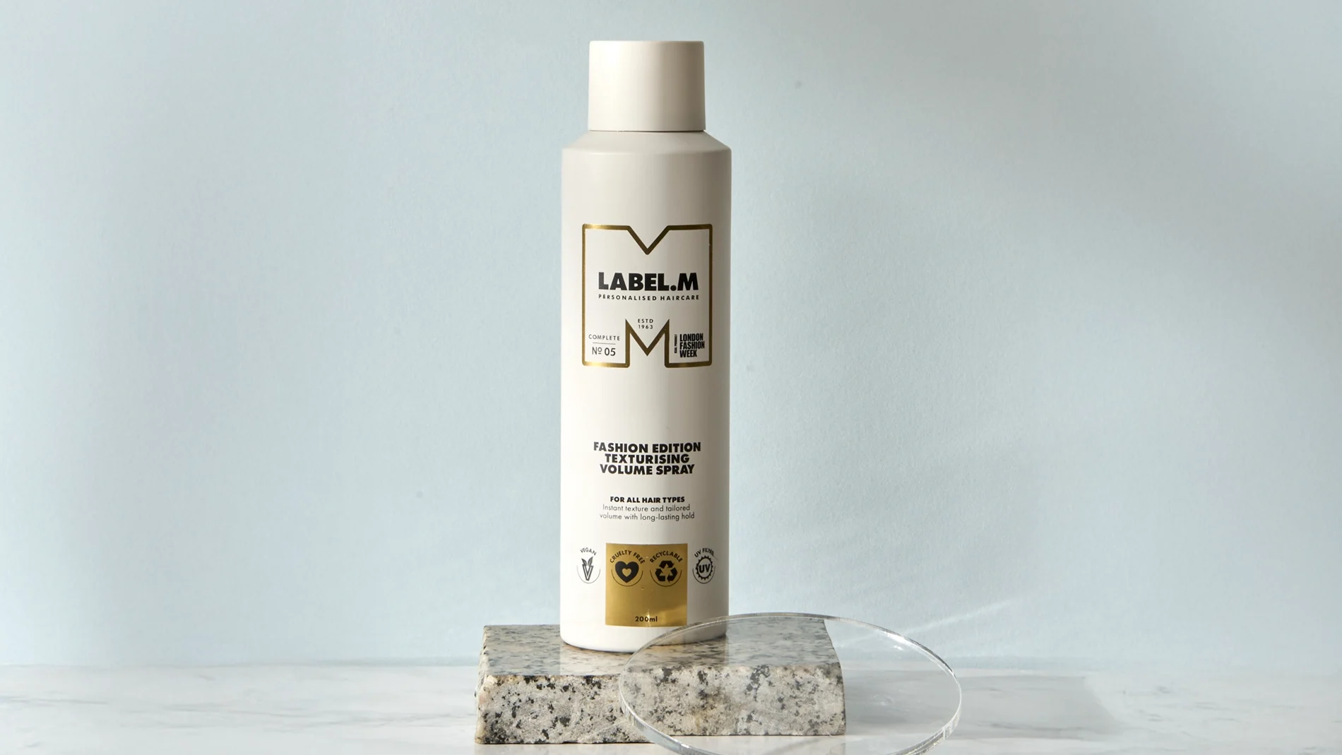 LABEL.M Fashion Edition Texturising Volume Spray i vit flaska placerad på ett stenblock med en transparent akrylskiva framför. Förpackningen visar texten “LABEL.M Personalised Haircare”, “Fashion Edition Texturising Volume Spray”, “For All Hair Types” och volymen 200 ml.